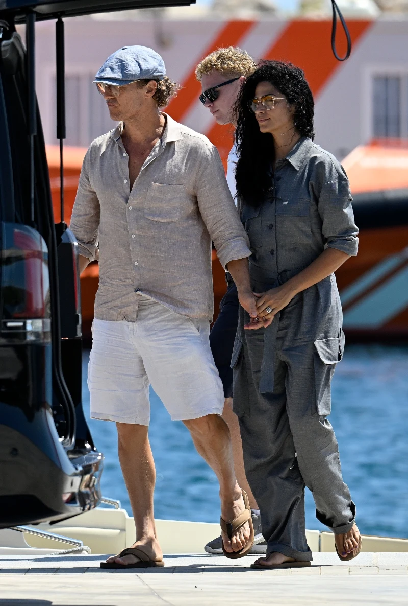 Matthew McConaughey i Camila Alves Para trzymająca się za ręce, ubrana w letnie, luźne ubrania spaceruje nad wodą, w tle widoczna łódź i odbijające się słońce.