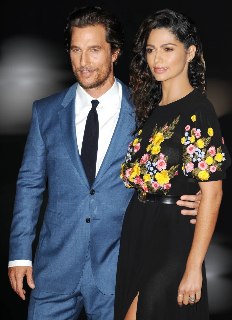 Matthew McConaughey i Camila Alves Elegancko ubrany mężczyzna w niebieskim garniturze z białą koszulą i czarnym krawatem stoi obok kobiety w czarnej sukience ozdobionej kolorowymi kwiatami, z ręką opartą na jej talii.