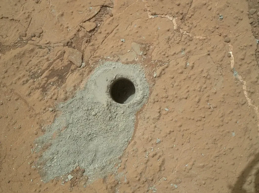 Curiosity w badanych próbkach z Marsa znalazł m.in. węgiel organiczny. Okrągły otwór wywiercony w jasnobrązowej, pylisto-kamiennej powierzchni z wyraźnie widocznym osadem pyłu wokół miejsca wiercenia.
