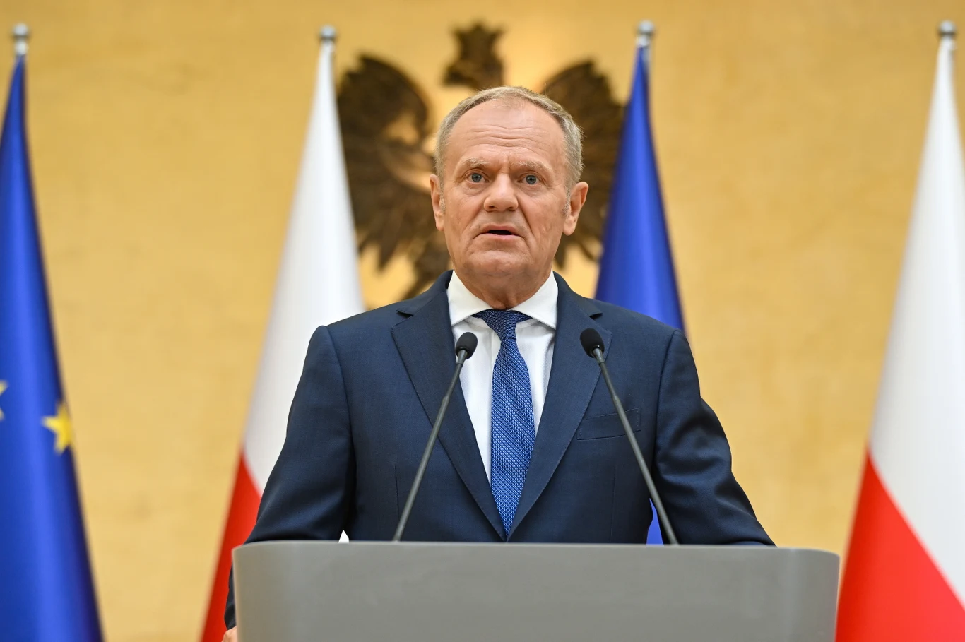 Premier Tusk zapewnia, że rząd jest gotowy do interwencji na rynku paliw Mężczyzna w garniturze przemawia przy mównicy, w tle widoczne flagi Polski i Unii Europejskiej oraz orzeł na ścianie.