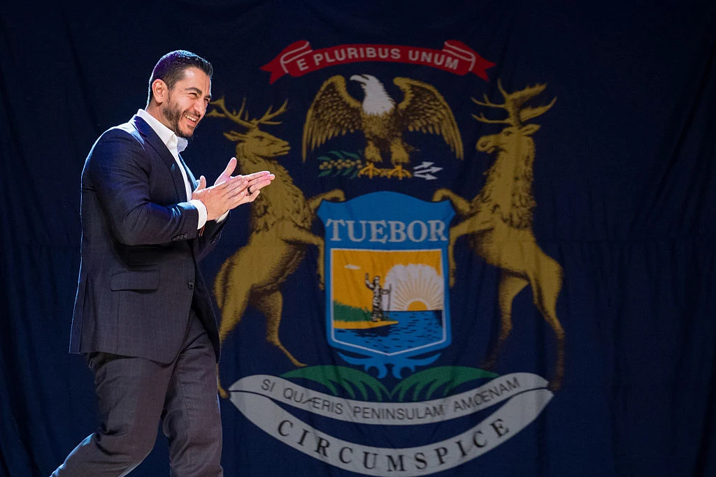 Abdul El-Sayed Mężczyzna w garniturze uśmiecha się i klaszcze, idąc przed dużym tłem z herbem stanu Michigan.