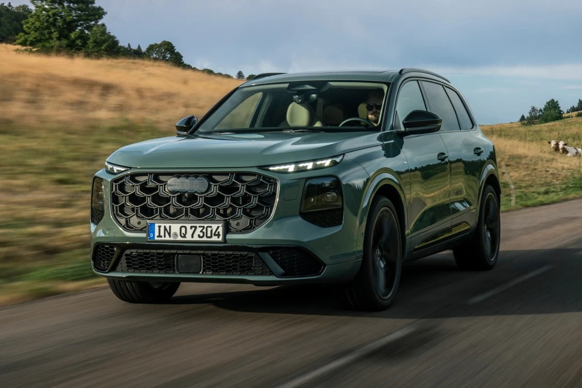 Jasnozielony SUV marki Audi z charakterystycznym, dużym grillem i nowoczesnymi reflektorami jadący po asfaltowej drodze w otoczeniu letniego krajobrazu z suchą trawą.