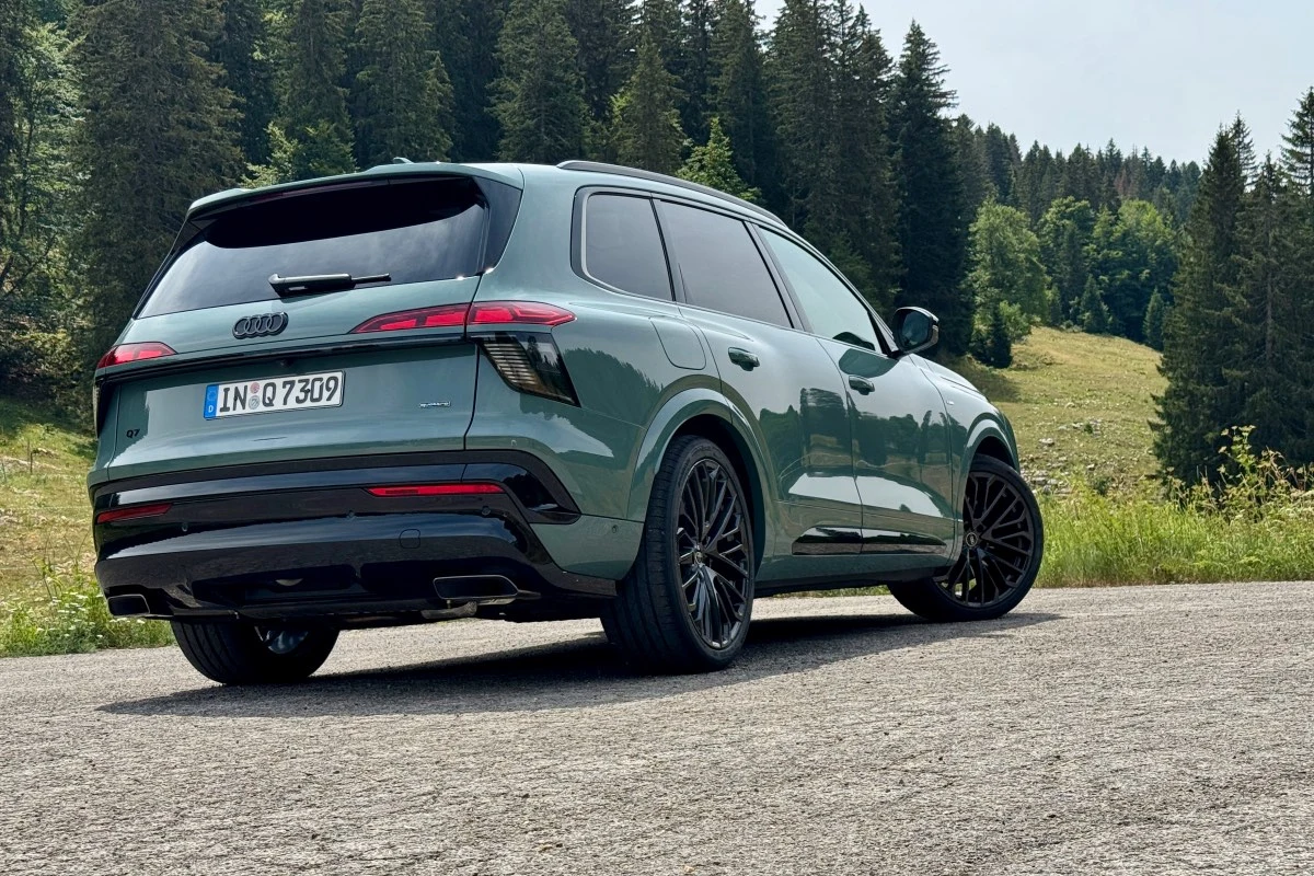 Zielony SUV marki Audi z czarnymi felgami i przyciemnianymi szybami stoi na asfaltowej drodze w otoczeniu lasu iglastego i pagórkowatego terenu.