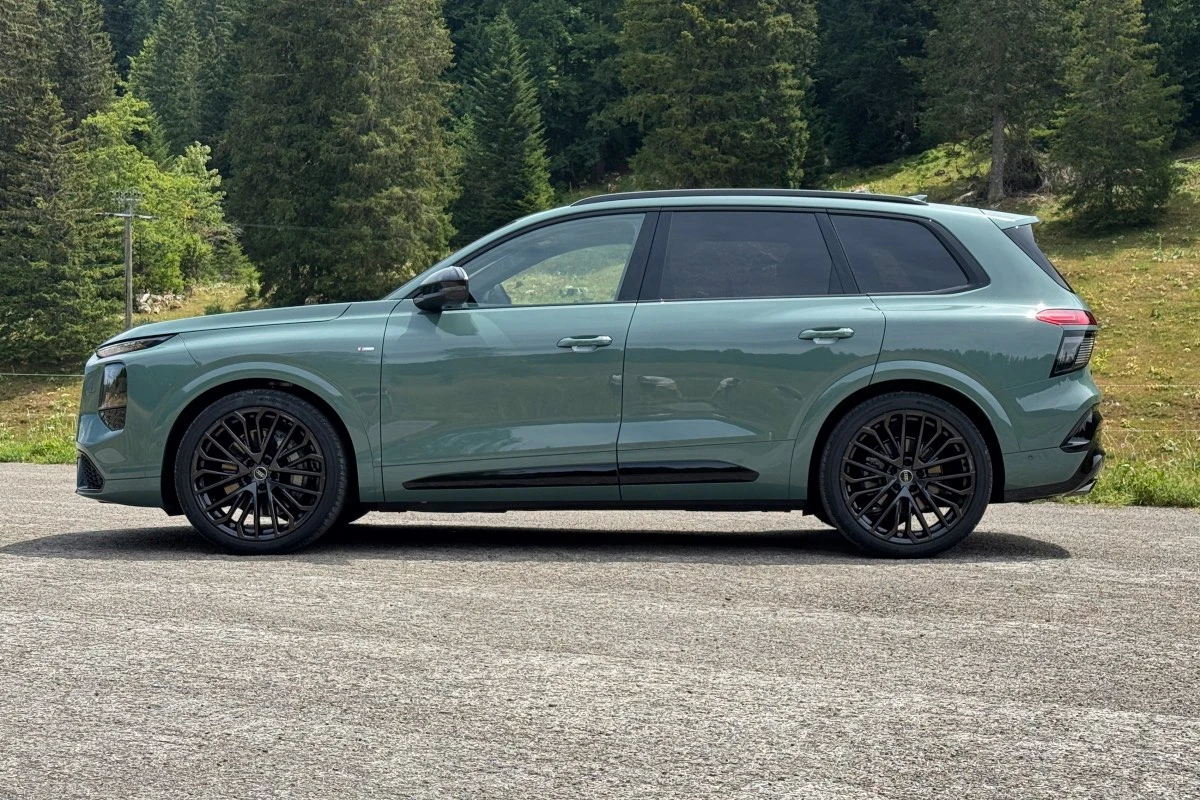 Jasnozielony SUV o nowoczesnej sylwetce na czarnych, wieloramiennych felgach stoi na asfaltowej nawierzchni z lasem i zielonym wzgórzem w tle.