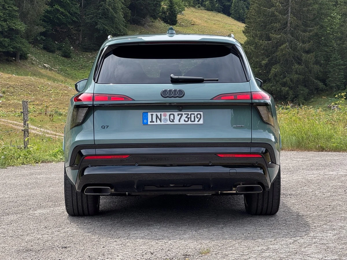 Tył zielonego Audi Q7 z widocznymi tylnymi światłami LED, podwójnymi końcówkami wydechu i niemiecką rejestracją, samochód stoi na asfaltowej drodze pośród zielonych łąk i drzew.