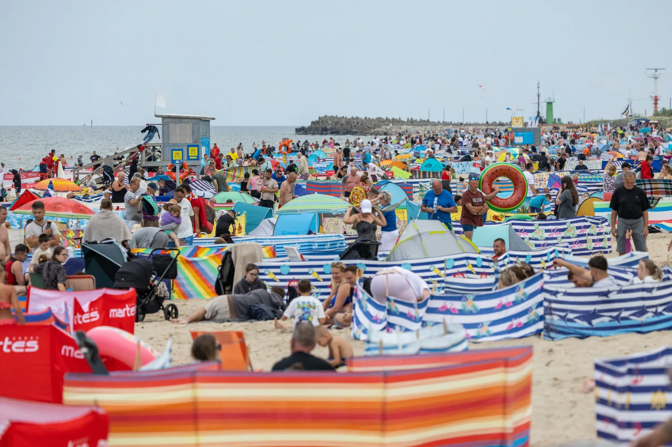 Rezerwacja miejsc na plaży i przy hotelowych basenach to wakacyjne hobby turystów Plaża z tłumem turystów, kolorowe parawany, parasole i leżaki, w tle morze oraz nadmorska infrastruktura.