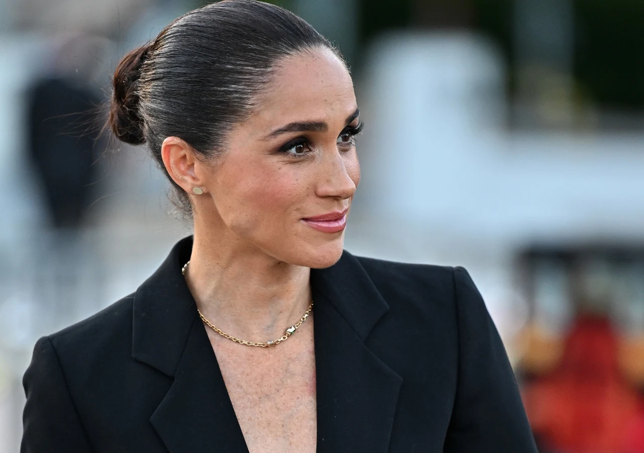 Meghan Markle Meghan Markle w czarnej marynarce i złotym naszyjniku, patrzy z uśmiechem na kogoś poza kadrem.
