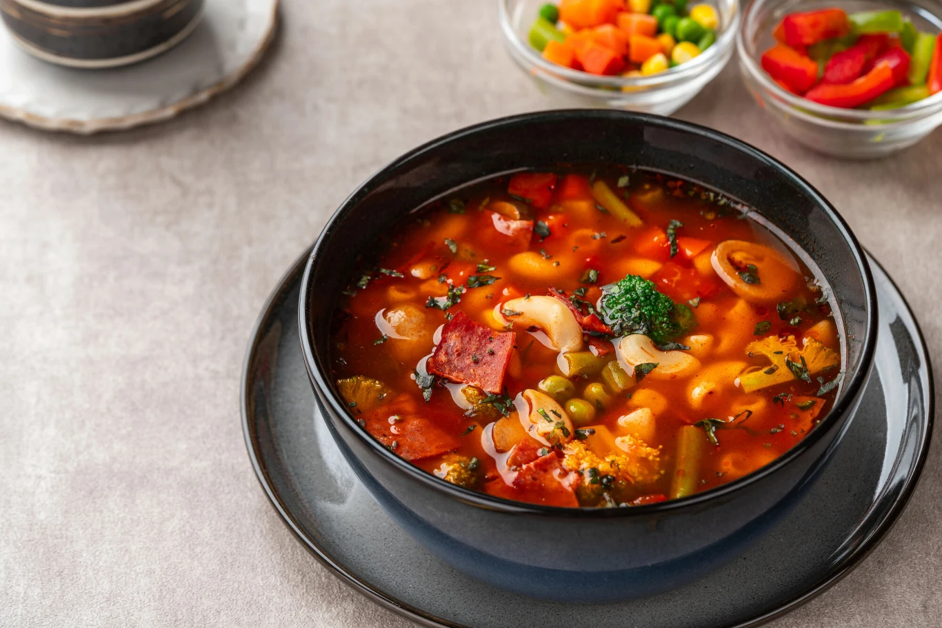 Menu w restauracji potrafi zmylić. Te "wykwintne" potrawy są bardzo proste Gorąca zupa z makaronem rurki, kawałkami kiełbasy, brokułem, groszkiem i marchewką podana w ciemnej misce na talerzu, w tle kolorowe miseczki z warzywami.