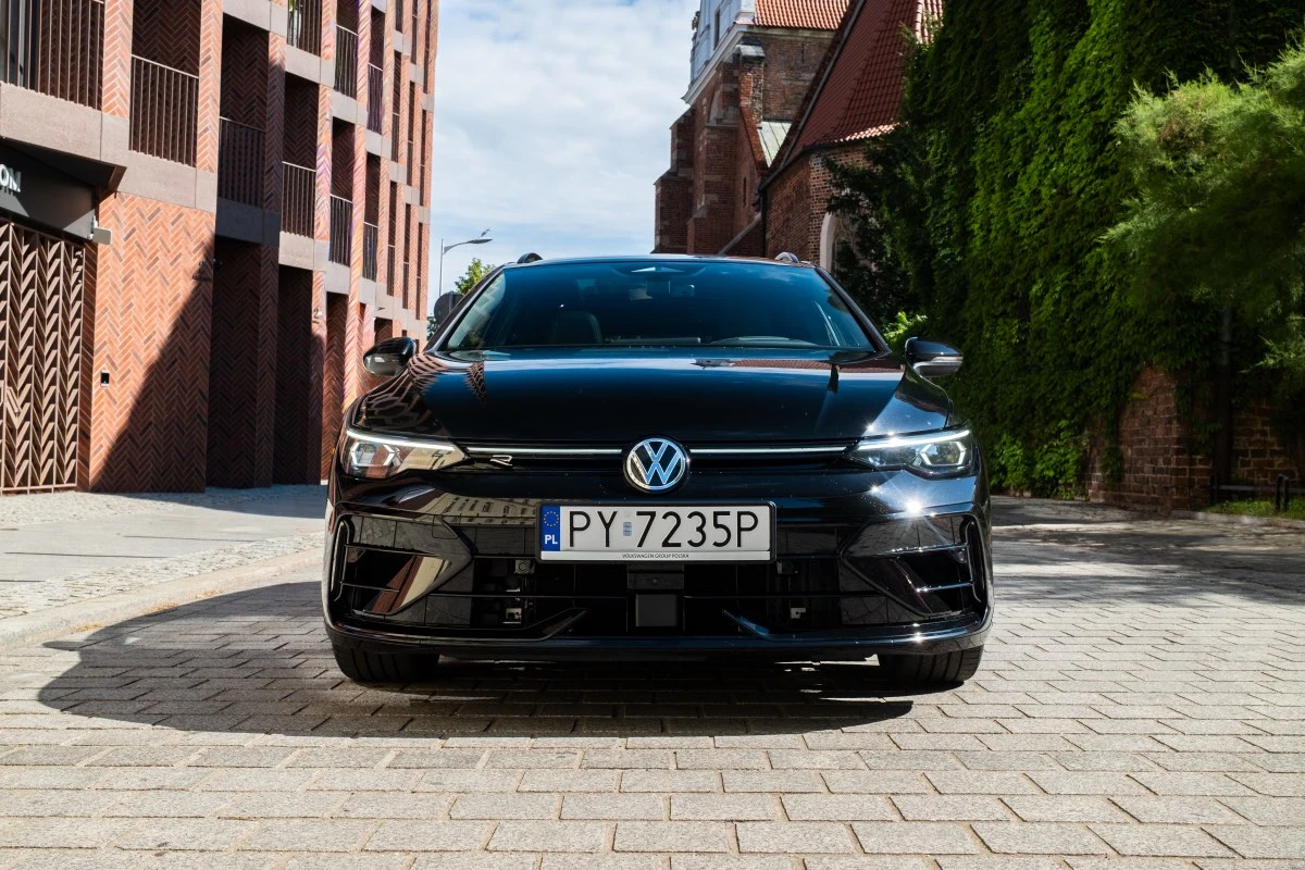 Volkswagen Golf R Czarny Volkswagen Golf R stoi zaparkowany przodem do aparatu na brukowanej ulicy, w otoczeniu nowoczesnych i historycznych budynków.