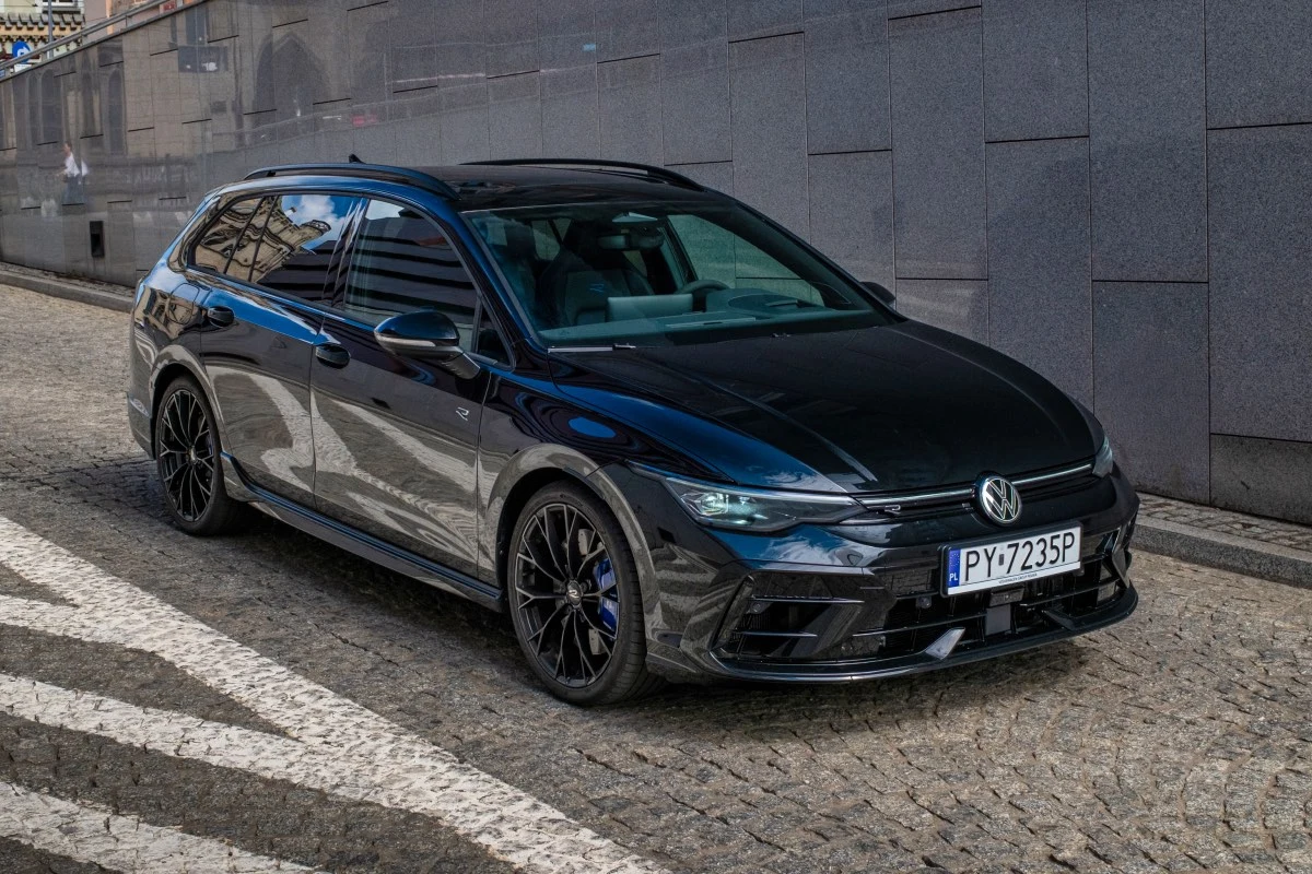 Czarny lakier sprawia, że Golf R wygląda jeszcze bardziej niepozornie. Czarny samochód marki Volkswagen zaparkowany na brukowanej ulicy przy szarym murze, z widocznymi czarnymi felgami i sportowym nadwoziem w wersji kombi.