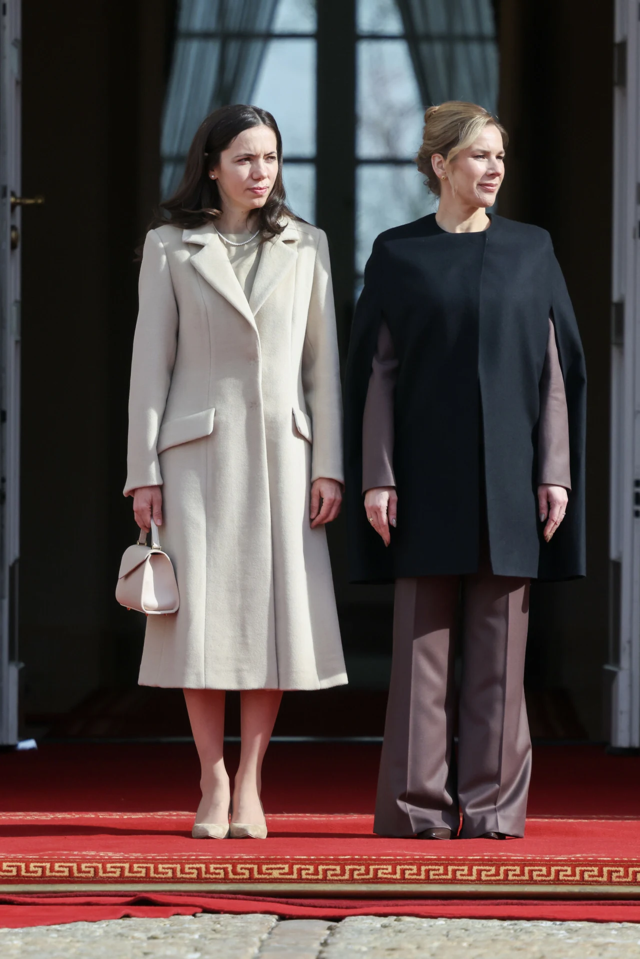 Marta Nawrocka nie boi się nieoczywistych połączeń w modzie Dwie elegancko ubrane kobiety stojące obok siebie na czerwonym dywanie przed wejściem do budynku, jedna w jasnym, długim płaszczu z małą torebką, druga w czarnym narzutce i beżowym zestawie ze spodniami.