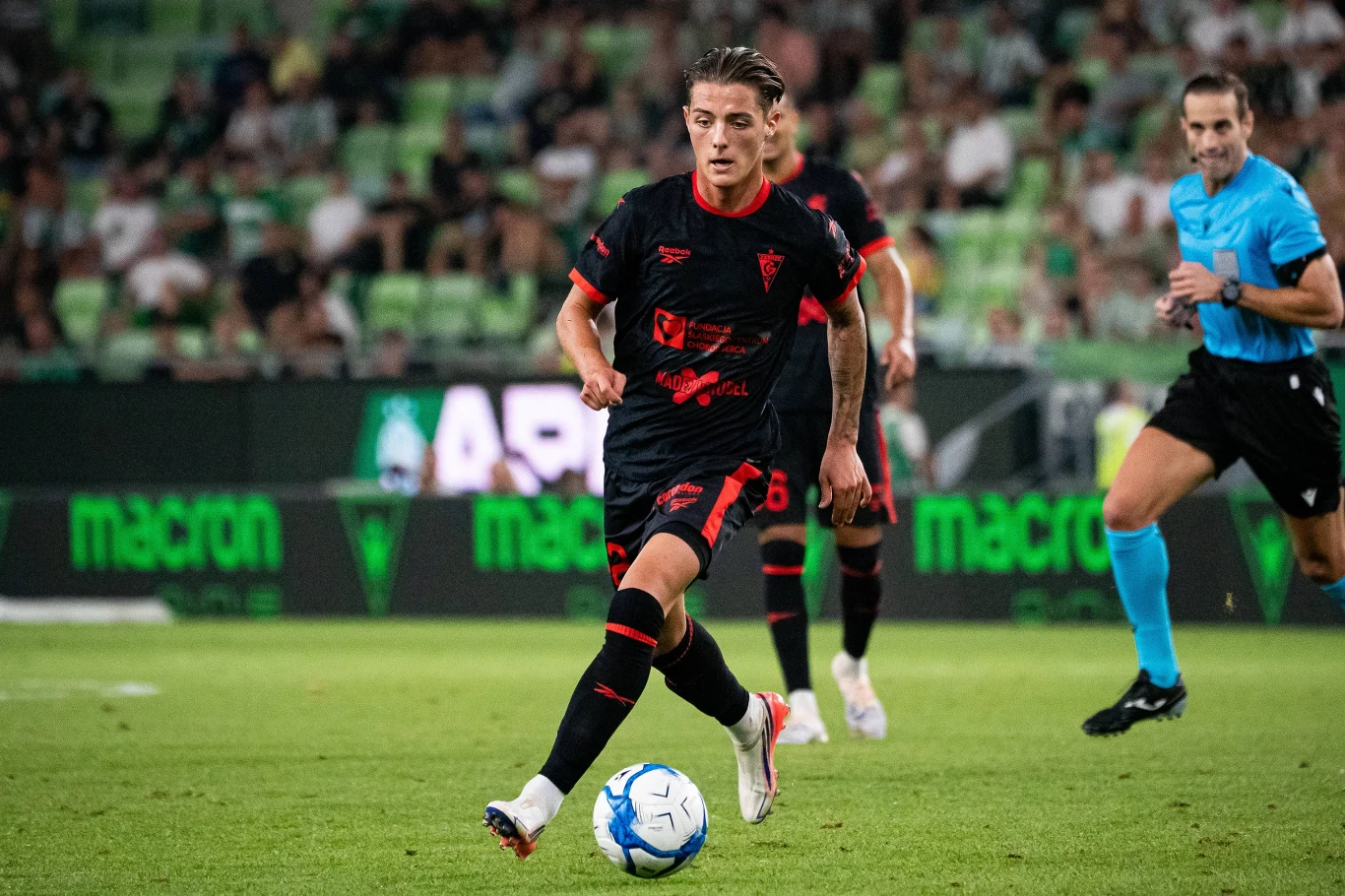 Kacper Urbański w barwach Górnika Zabrze Kacper Urbański z Górnika Zabrze w czarno-czerwonym stroju prowadzi piłkę podczas meczu, wokół inni piłkarze i sędzia.