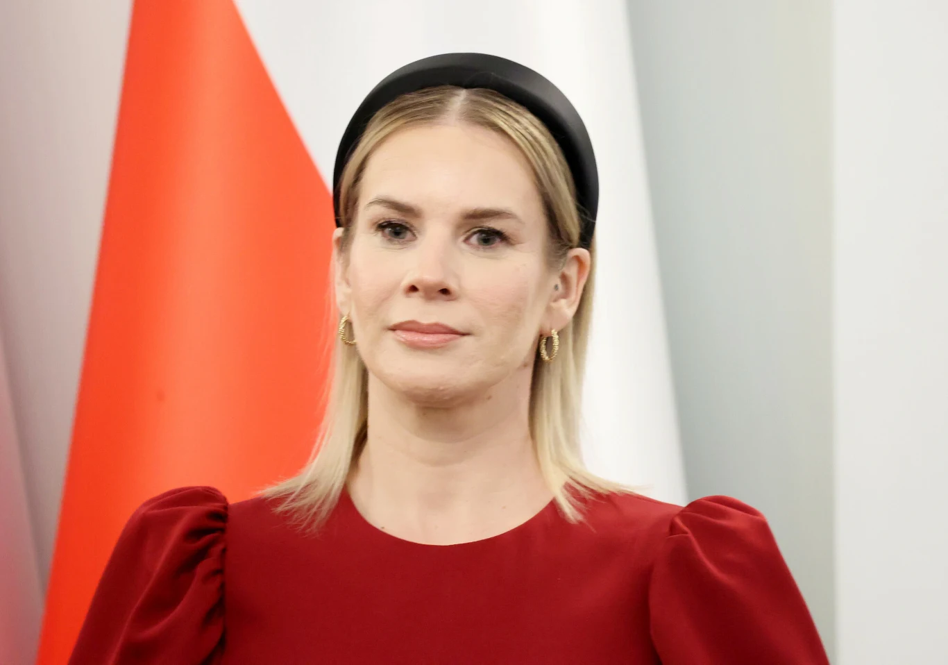 Jak Polacy oceniają pierwszy rok pełnienia roli pierwszej damy przez Martę Nawrocką? Kobieta o blond włosach ubrana w bordową sukienkę z bufiastymi rękawami, na głowie czarna opaska, w tle fragment biało-czerwonej flagi.