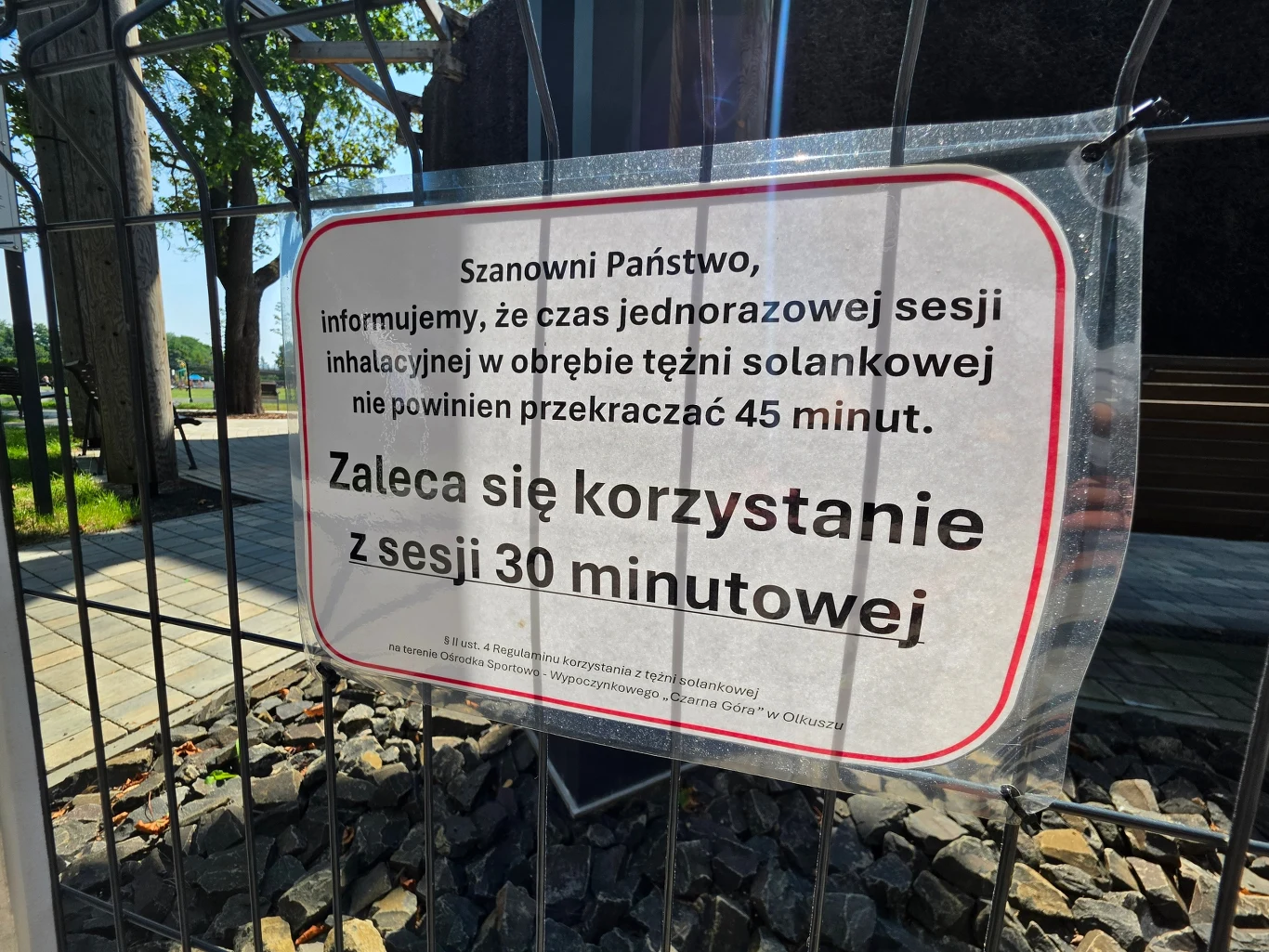 Informacyjna tabliczka przy ogrodzeniu z zaleceniem maksymalnego czasu korzystania z sesji inhalacyjnej w tężni solankowej, sugerująca 30-minutowe sesje.