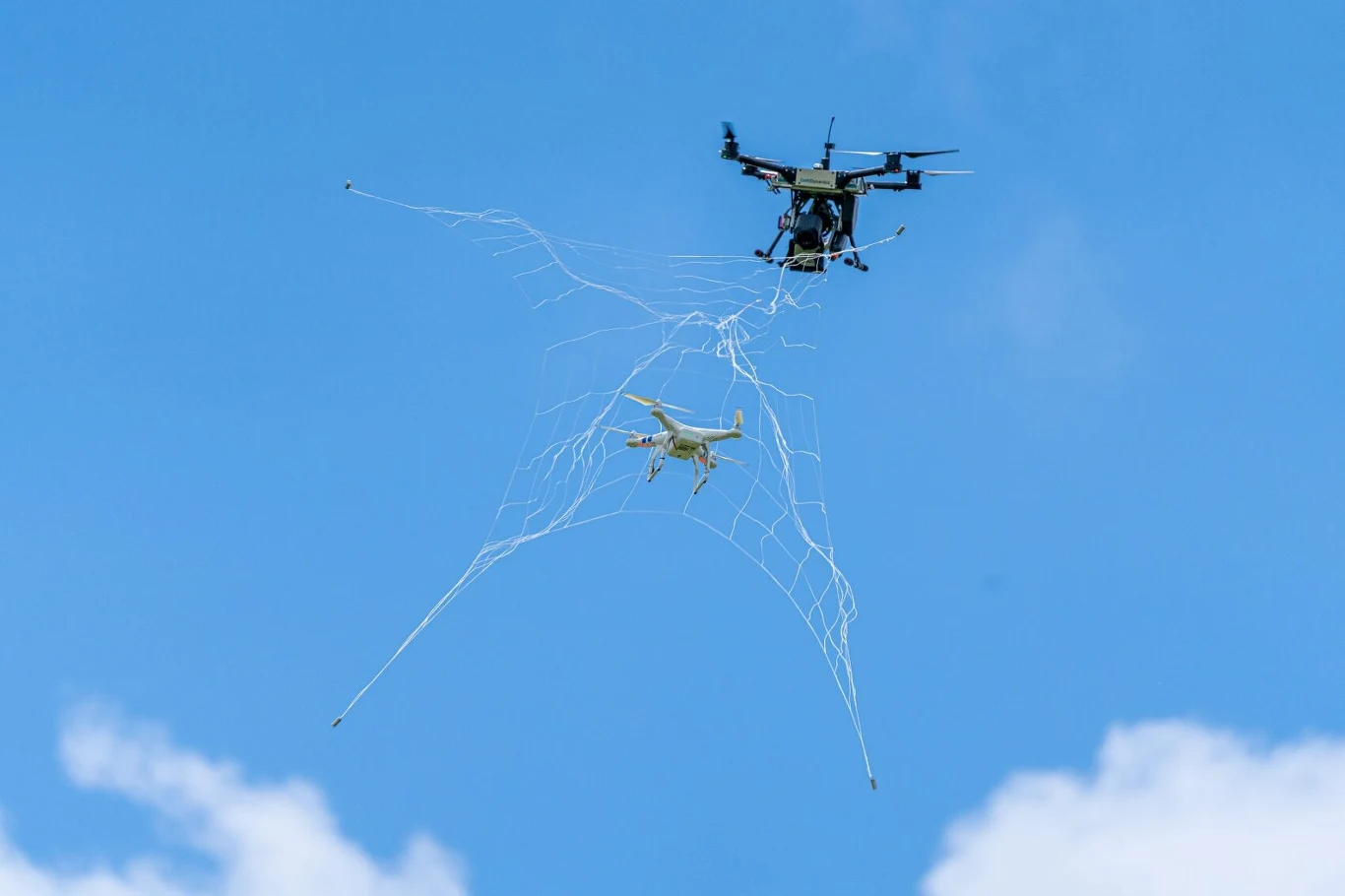 Duży dron wyposażony w sieć łapie mniejszego drona unoszącego się w powietrzu na tle jasnego, niebieskiego nieba.