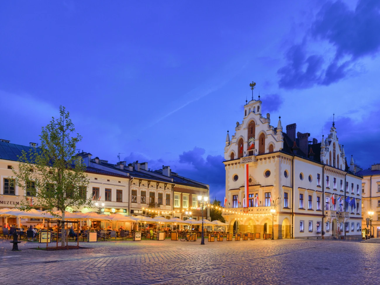 Jak przetrwać noce tropikalne w Polsce? Wieczorny rynek z zabytkowym ratuszem, oświetlony, lokale gastronomiczne i goście pod parasolami.