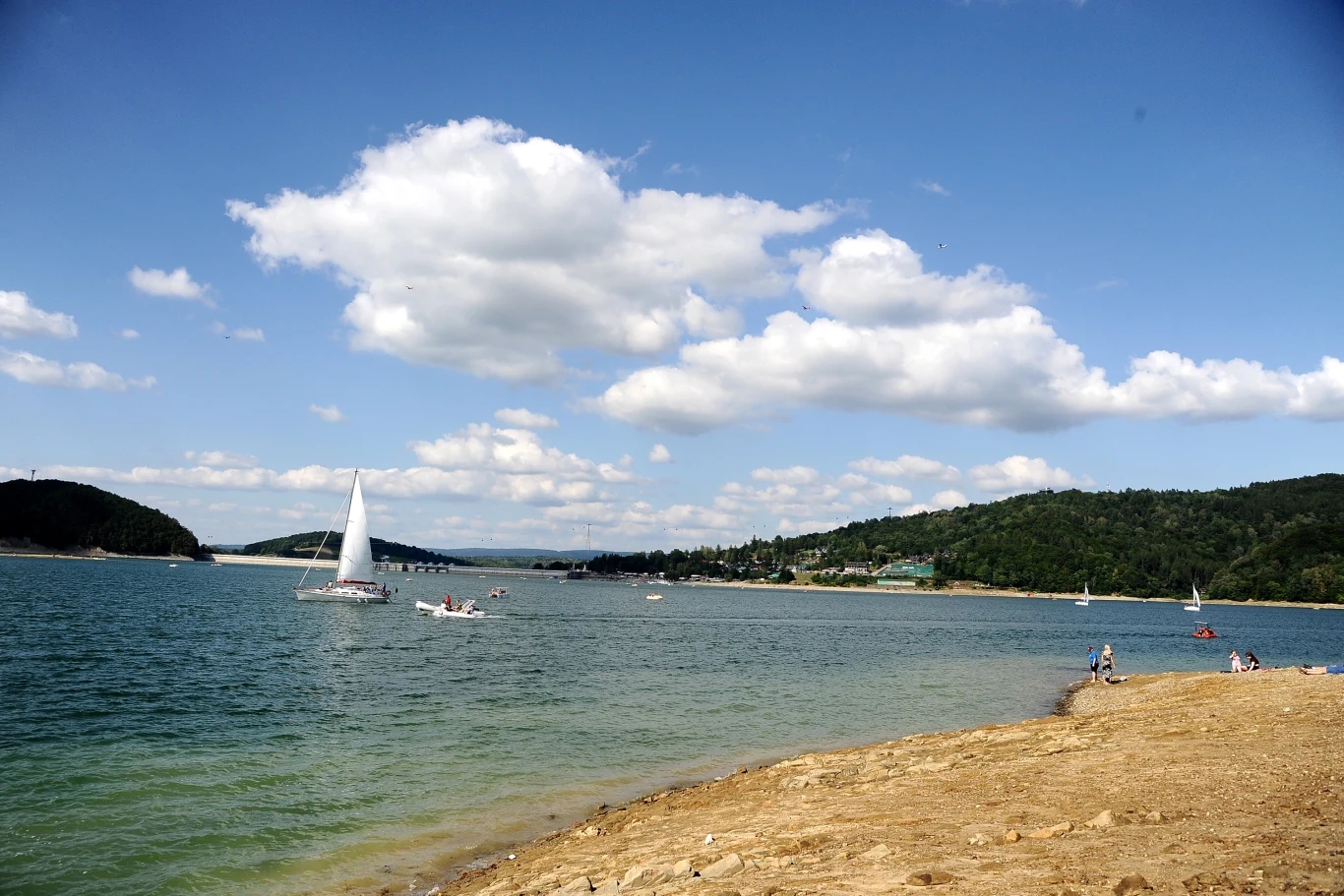 Bieszczadzkie morze to idealna alternatywa dla Bałtyku Jacht żaglowy na Jeziorze Solińskim, osoby na plaży, zielone wzgórza i niebo z białymi chmurami w tle.