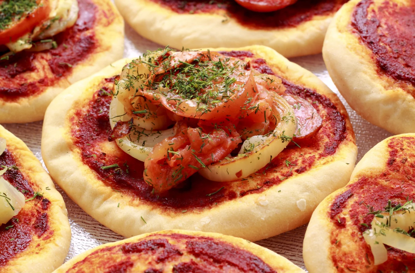 Pizzerinki to idealna przekąska na imprezę Małe, okrągłe pizzerinki z puszystym ciastem, sosem pomidorowym, cebulą, pomidorami i ziołami na blasze.