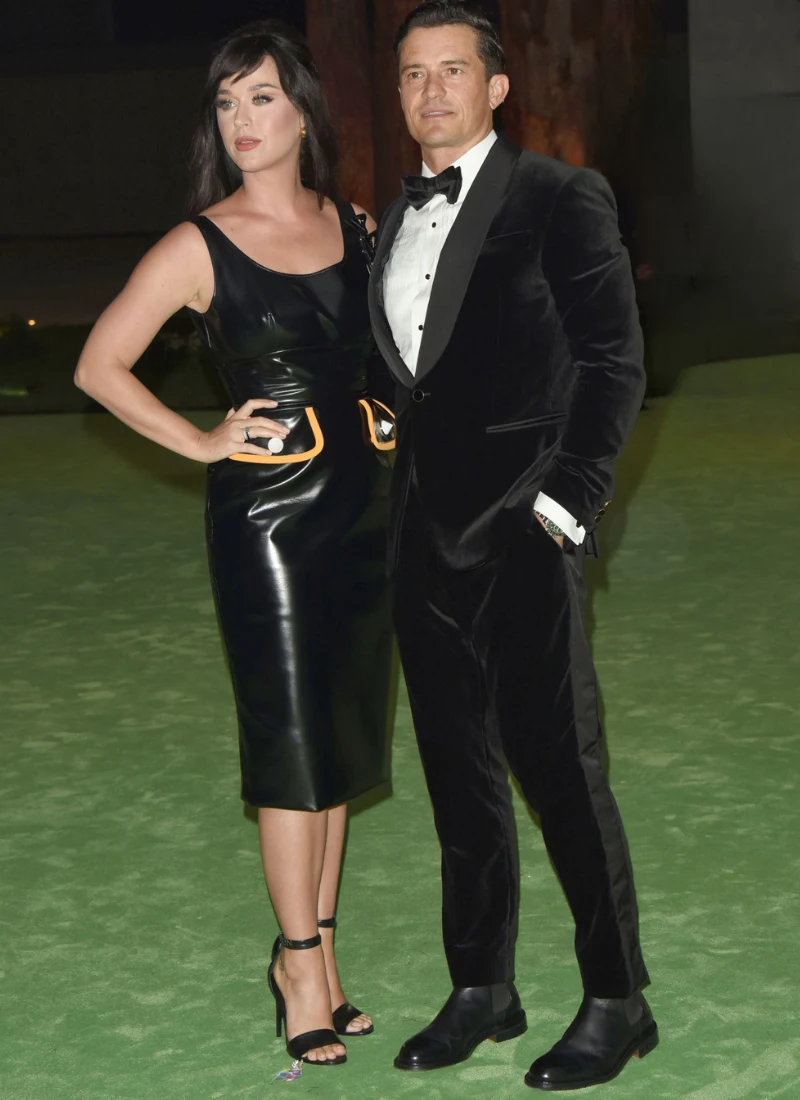 Orlando Bloom i Katy Perry Elegancka kobieta w czarnej, błyszczącej sukience midi i czarnych wysokich sandałach stoi obok mężczyzny w czarnym, aksamitnym smokingu i muchą, oboje pozują razem na zielonym dywanie.
