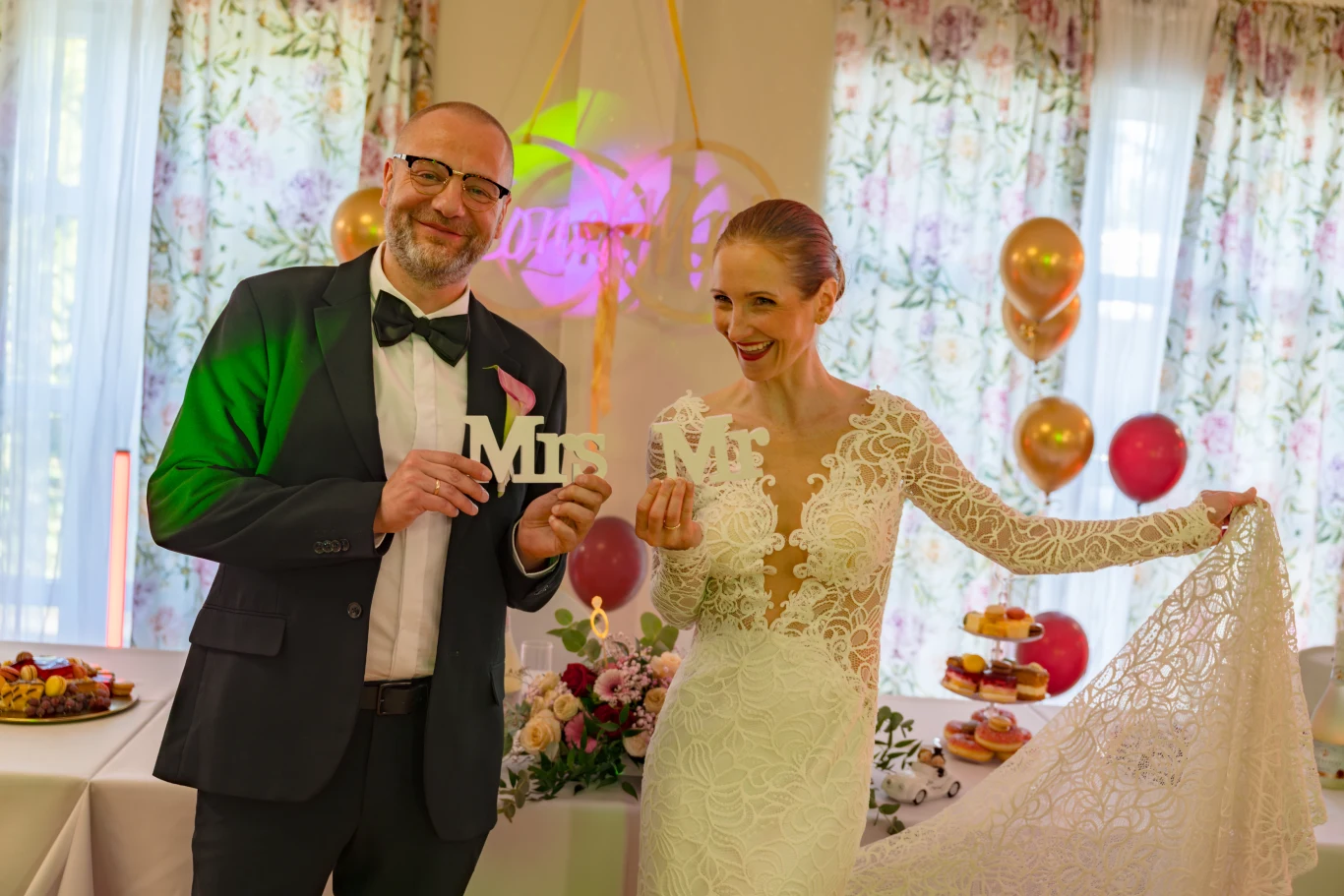 Kadr z serialu "Sprawiedliwi – Wydział Kryminalny" Para młoda w eleganckich strojach trzyma napisy „Mr” i „Mrs” w ozdobnej sali weselnej z balonami i stołem pełnym słodkości w tle.