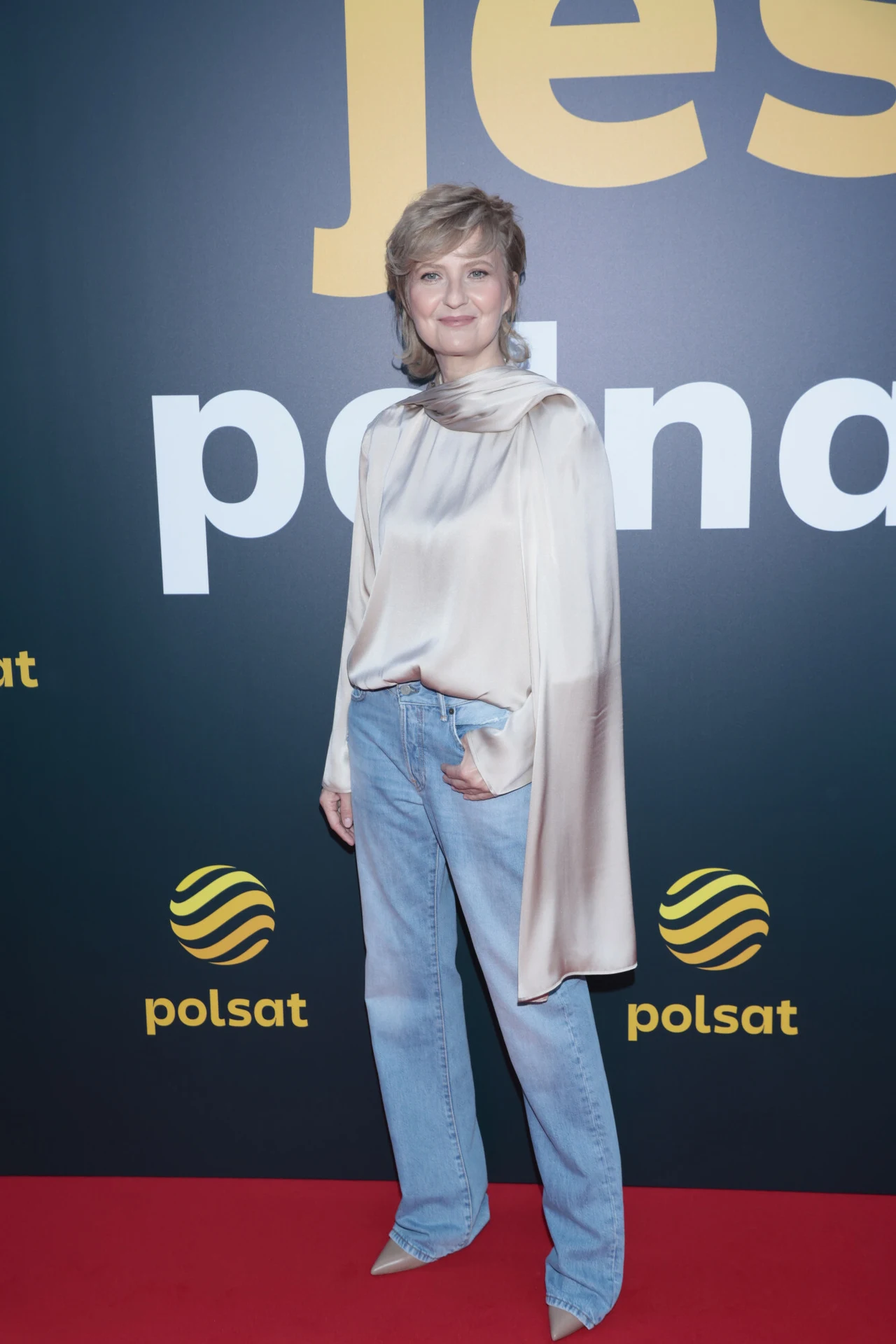 Kobieta w jasnej bluzce z długimi rozszerzanymi rękawami i niebieskich dżinsach, stojąca na czerwonym dywanie na tle ścianki z logotypami Polsatu.