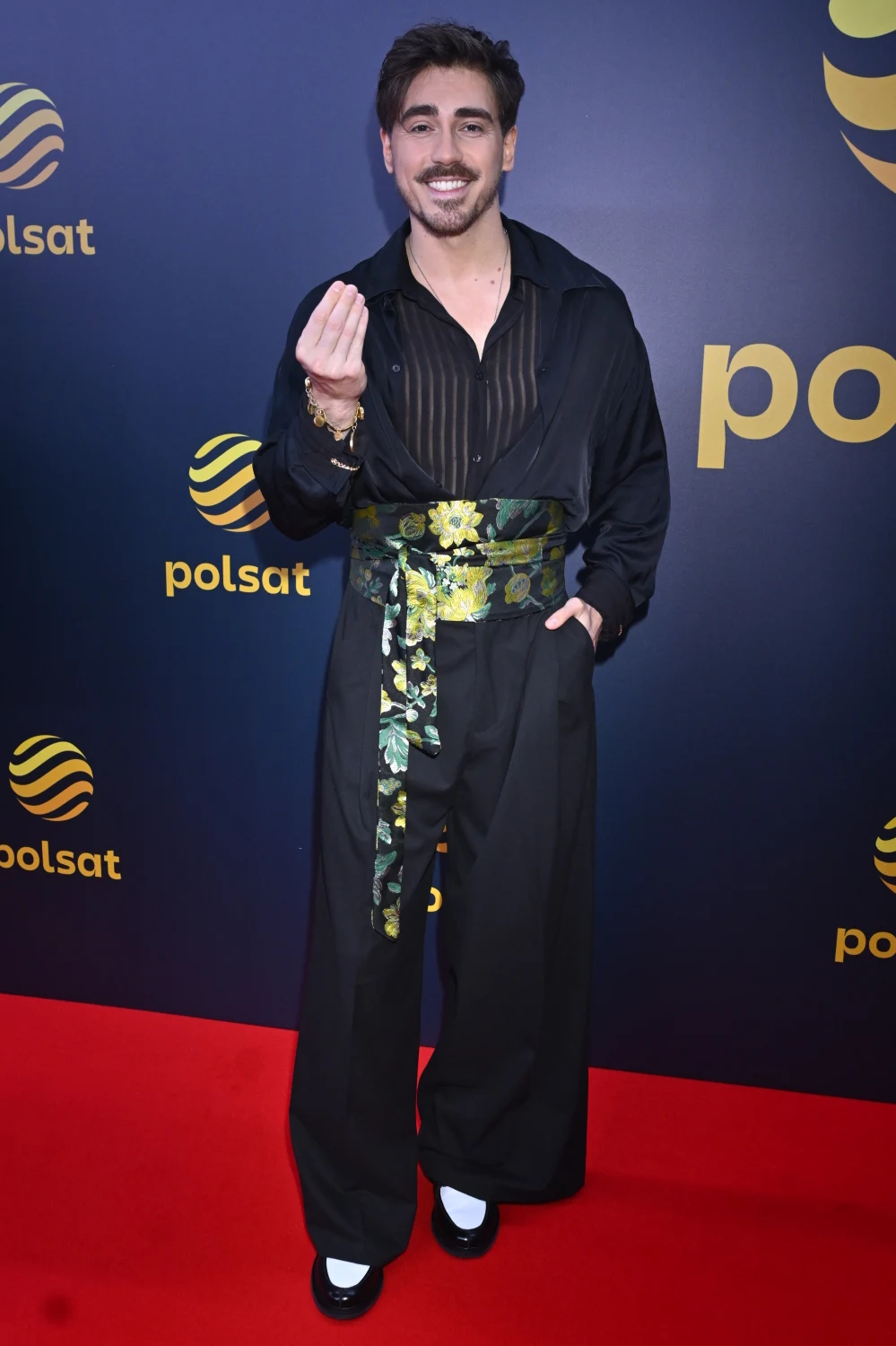 Mężczyzna z ciemnymi włosami ubrany w czarną koszulę z prześwitującymi pasami oraz szerokie, eleganckie spodnie z ozdobnym, błyszczącym pasem w kwiatowe wzory, stojący na czerwonym dywanie przed ścianką z logotypami Polsatu.