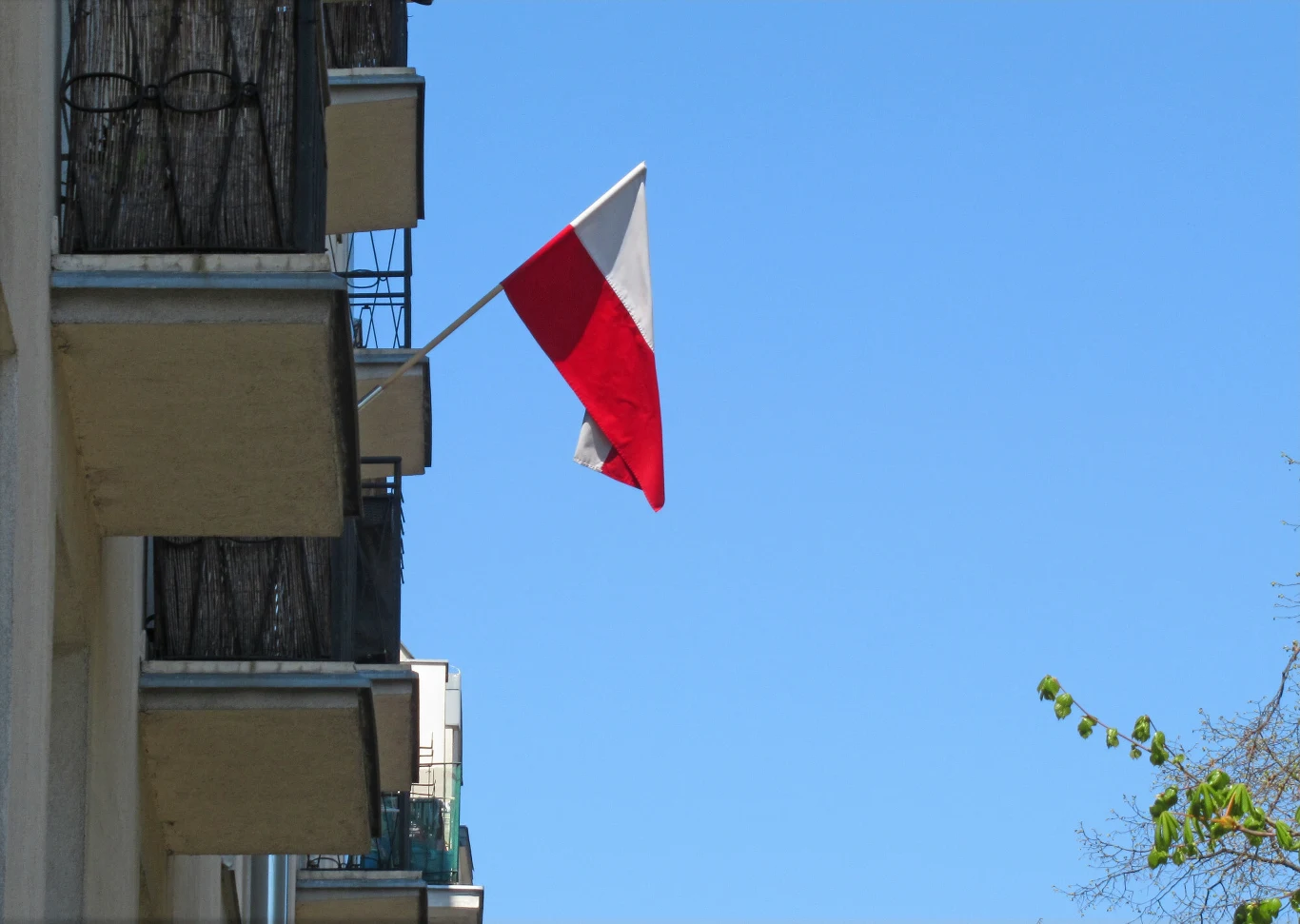 Polska flaga powiewająca na wysięgniku zamontowanym na balkonie bloku mieszkalnego, na tle czystego nieba.