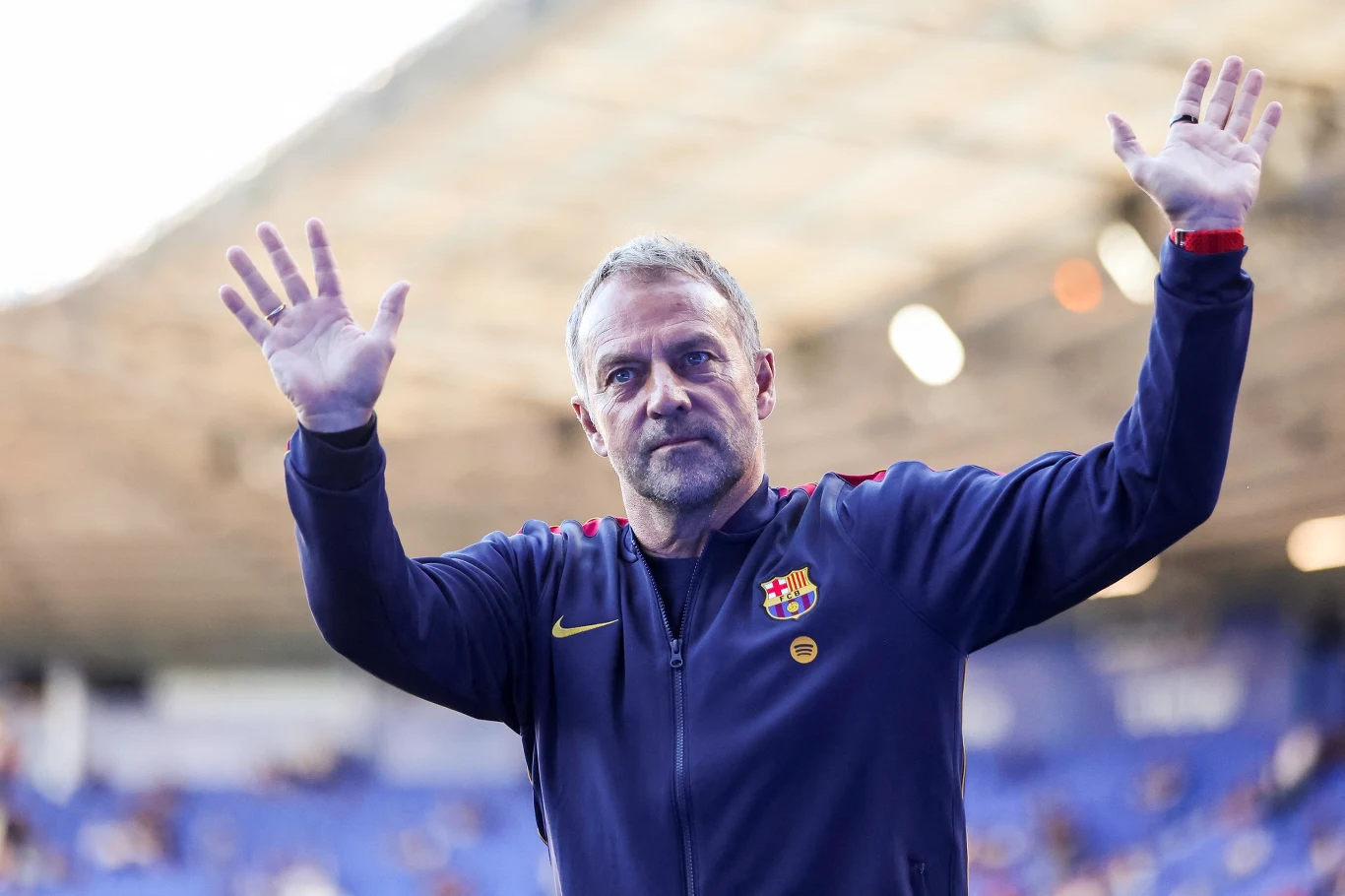 Mężczyzna w klubowej kurtce FC Barcelona z uniesionymi dłońmi stoi na tle stadionu.