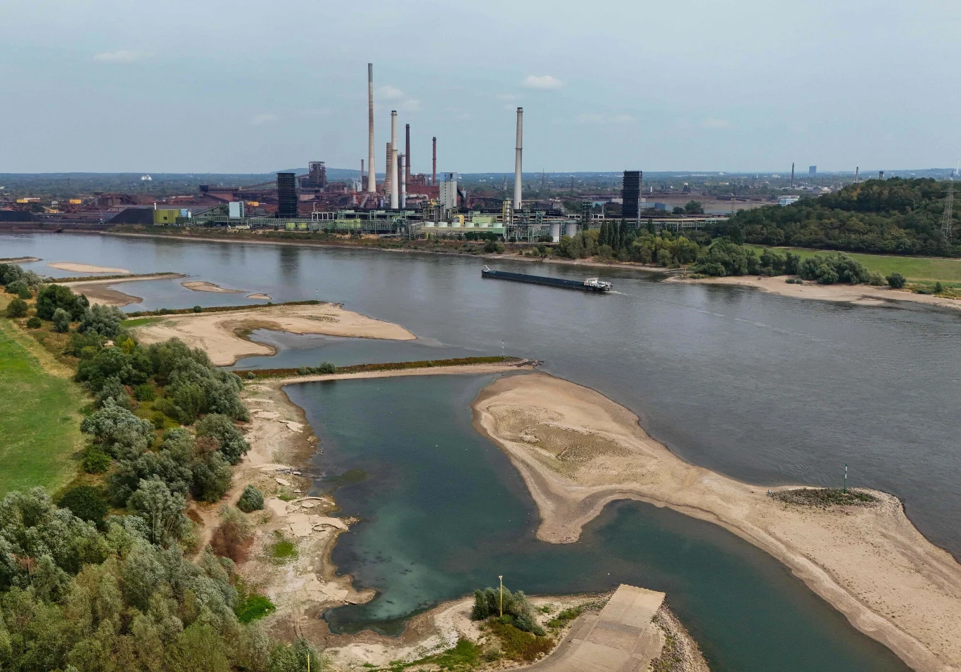 Niski stan wód w Niemczech negatywnie odbija się m.in. na transporcie stali i produktów chemicznych Rzeka z widocznymi odsłoniętymi piaszczystymi łachami, otoczona zielenią, w tle duży kompleks przemysłowy z wieloma wysokimi kominami.