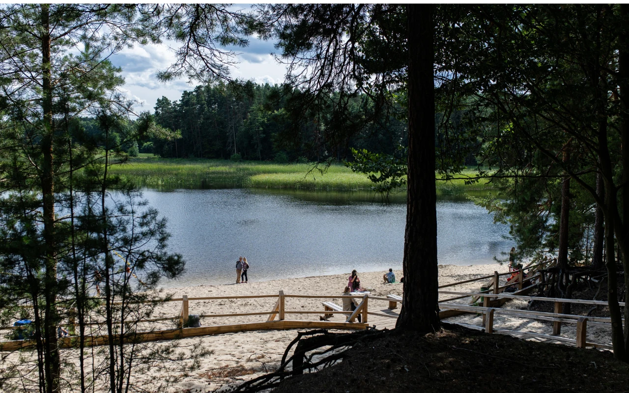 Piaszczysta plaża nad jeziorem otoczonym lasem, kilka osób spaceruje i odpoczywa w cieniu drzew, drewniane ogrodzenie oddziela ścieżkę od brzegu wody.