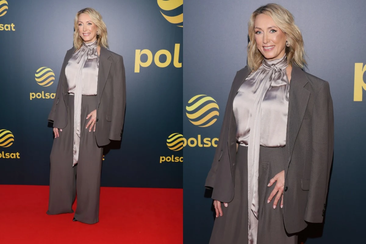 Kobieta w eleganckim, szarym garniturze i błyszczącej koszuli stoi na czerwonym dywanie na tle ścianki z logo Polsat, uśmiechając się do aparatu.