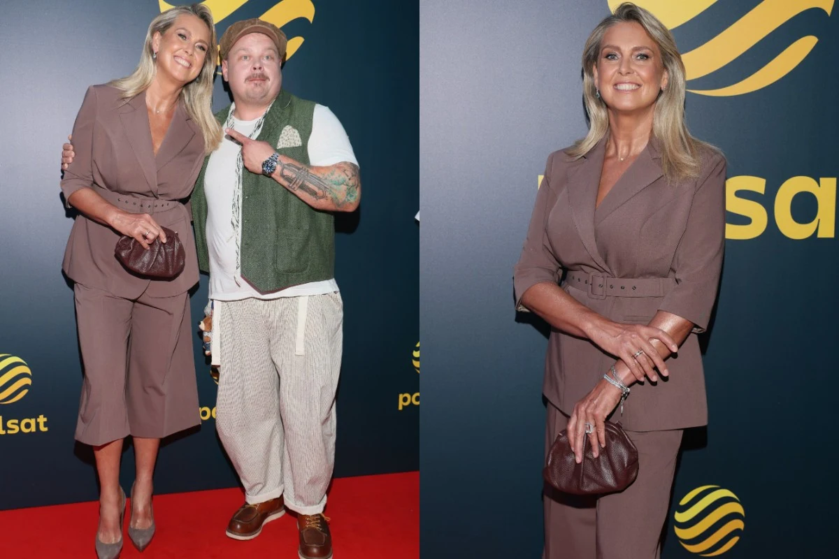 Kobieta w eleganckim garniturze w odcieniu brązu i jasnych szpilkach pozuje na ściance, trzymając ciemną kopertówkę, obok niej stoi mężczyzna w jasnych spodniach, białej koszulce i zielonej kamizelce, wskazując na towarzyszkę na tle logotypów Polsatu.