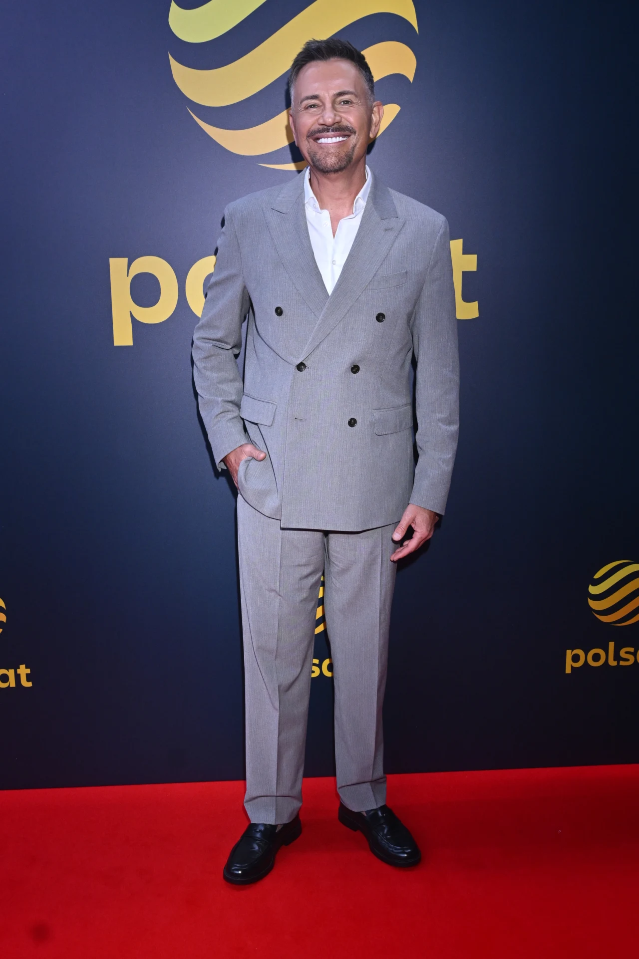 Mężczyzna w szarym garniturze i białej koszuli pozuje na czerwonym dywanie przed ścianą z logo Polsat.