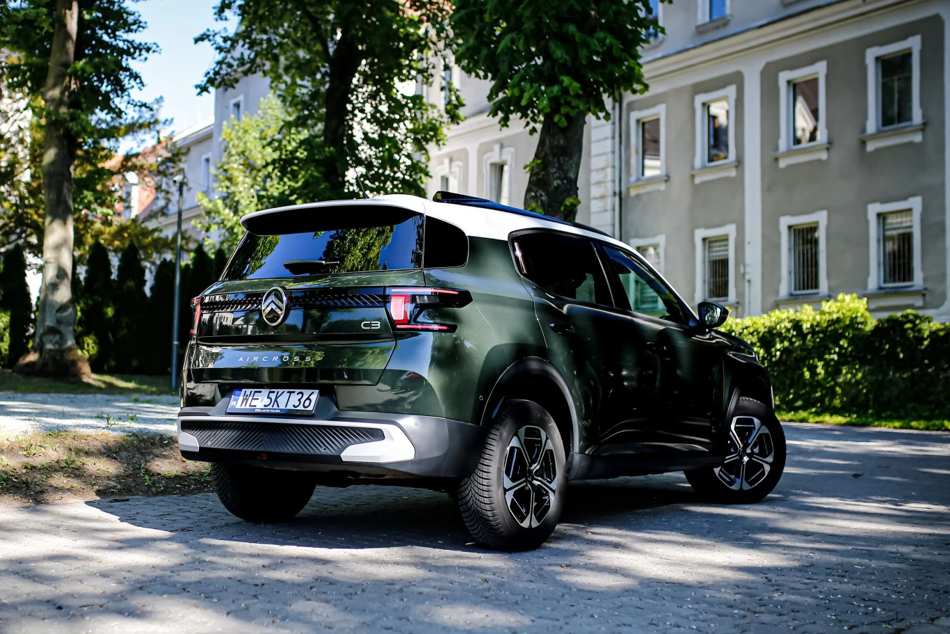 Ciemnozielony Citroën C5 Aircross zaparkowany na brukowanym parkingu, otoczony zielonymi drzewami i jasnymi budynkami mieszkalnymi w tle.