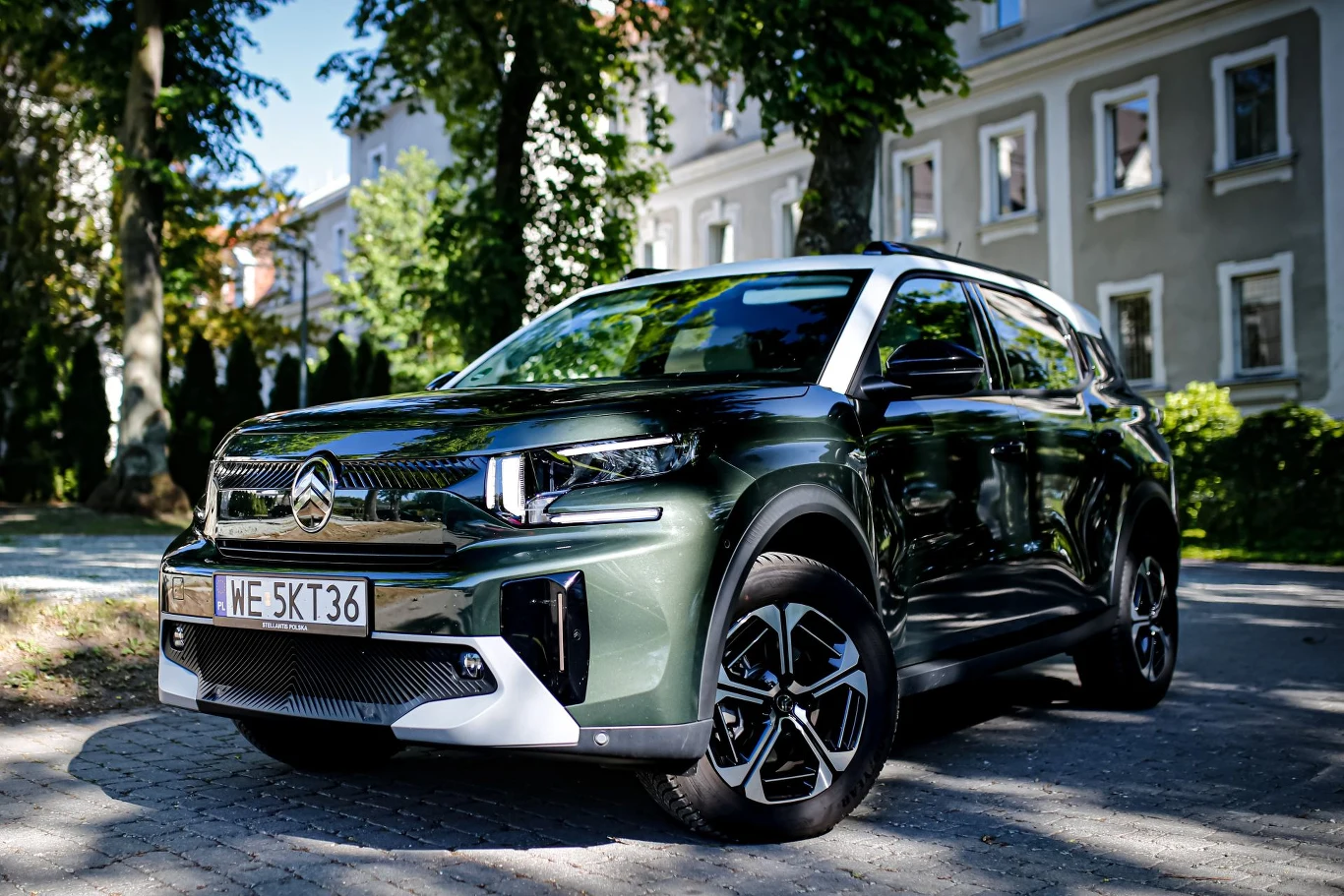Nowoczesny SUV w kolorze zielonym z czarnymi elementami nadwozia, stojący na brukowanej ulicy w otoczeniu zieleni i starych kamienic.
