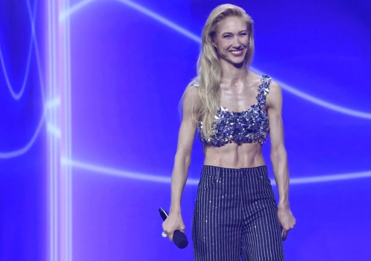 Ida Nowakowska na prezentacji jesiennej ramówki Polsatu Kobieta z długimi blond włosami w błyszczącym topie na ramiączkach i spodniach w paski, uśmiechnięta, trzyma mikrofon na tle niebieskiego, nowoczesnego oświetlenia scenicznego.