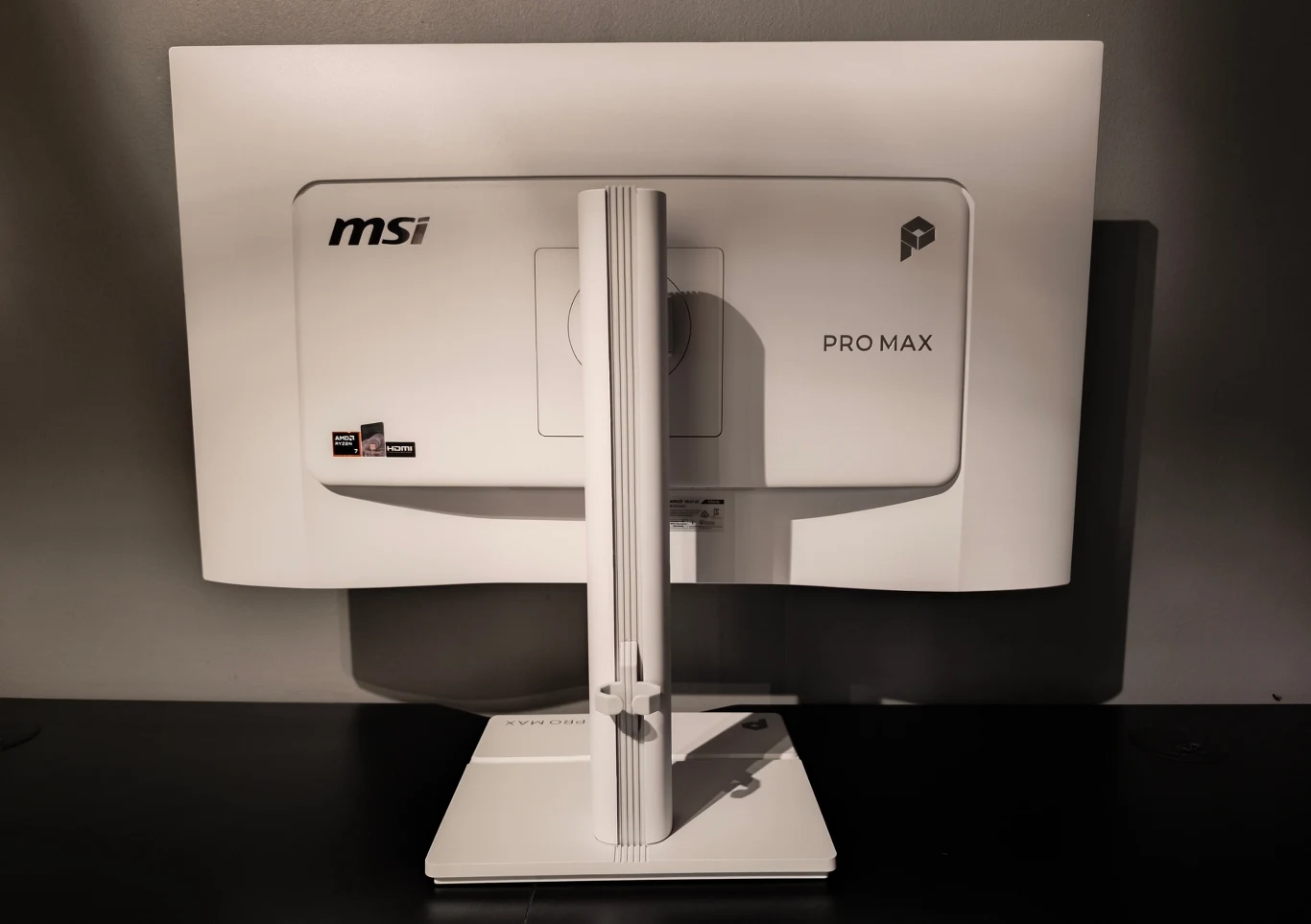 MSI PRO MAX 27P Z Biały monitor komputerowy marki MSI PRO MAX ustawiony na masywnej podstawie, widoczny z tyłu na tle ciemnej ściany.