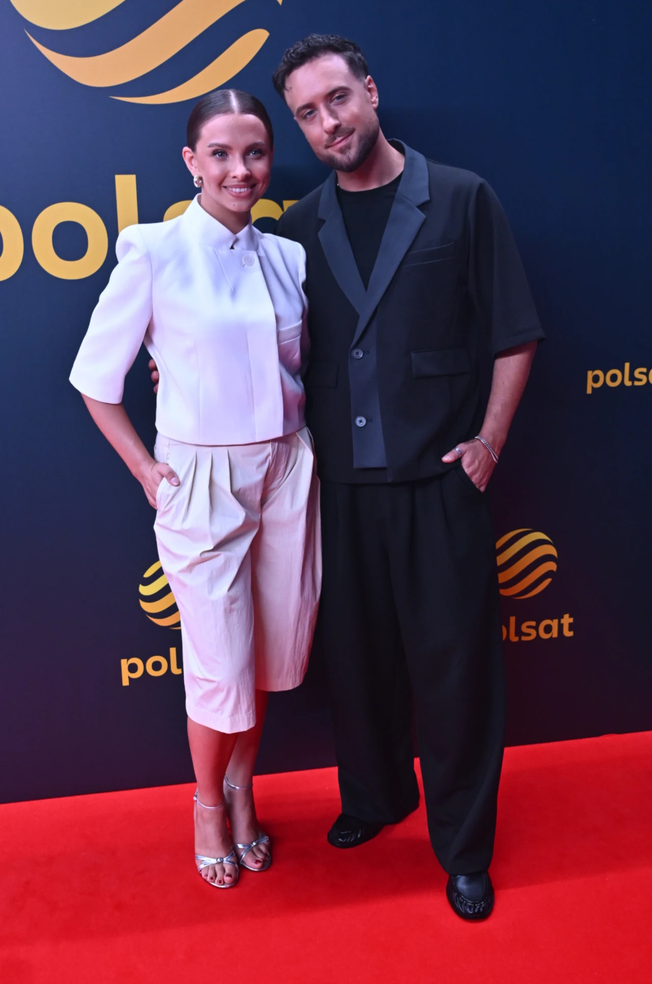 Ola Filipek i Aleksander Sikora na jesiennej ramówce Polsatu Para w eleganckich, nowoczesnych stylizacjach pozuje razem na czerwonym dywanie na tle ścianki z logo Polsat; kobieta w białym, zgeometryzowanym komplecie oraz srebrnych sandałkach, mężczyzna w czarnym, luźnym garniturze i sportowym obuwiu.