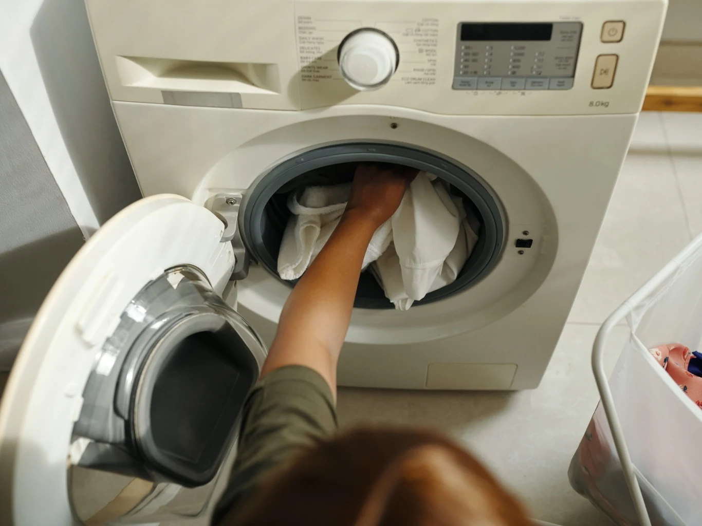 Gdy wnętrze pralki brzydko pachnie to znak, że trzeba ją wyczyścić Ręka wkłada białe pranie do otwartej pralki automatycznej, obok stoi kosz na brudne ubrania.