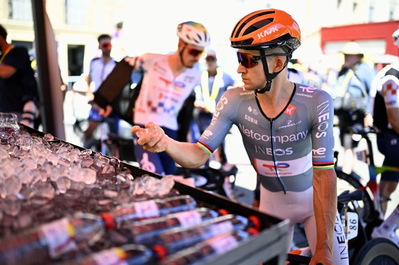 Michał Kwiatkowski Kolarz w stroju drużyny INEOS sięga po wodę z pojemnika wypełnionego lodem, wokół niego pozostali zawodnicy przygotowują się do jazdy na rowerach wyścigowych.