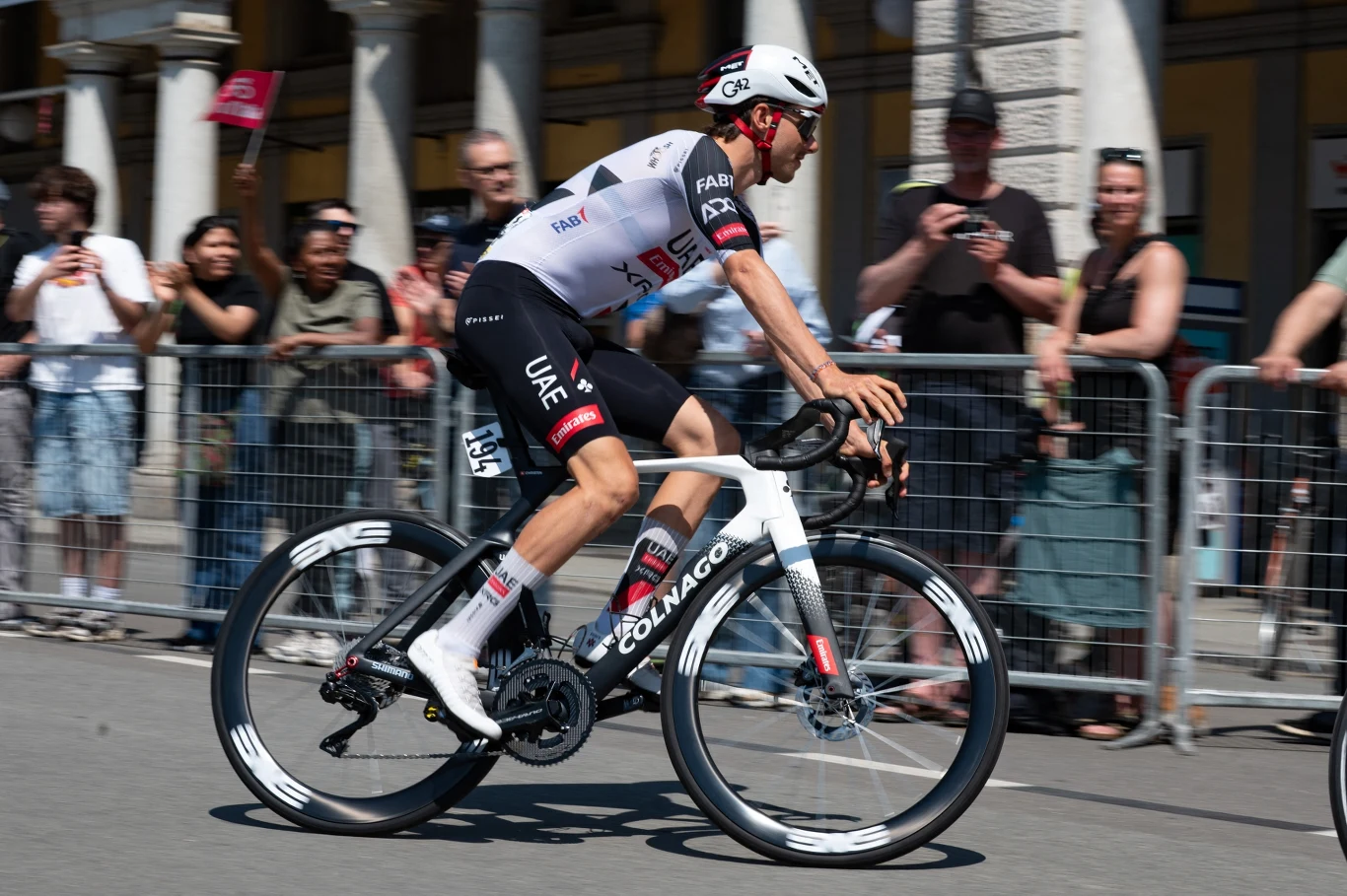 Jan Christen Zawodowy kolarz w stroju drużyny UAE Team Emirates jedzie na wyścigowym rowerze Colnago przy barierkach, wokół zgromadzona publiczność obserwuje wyścig.