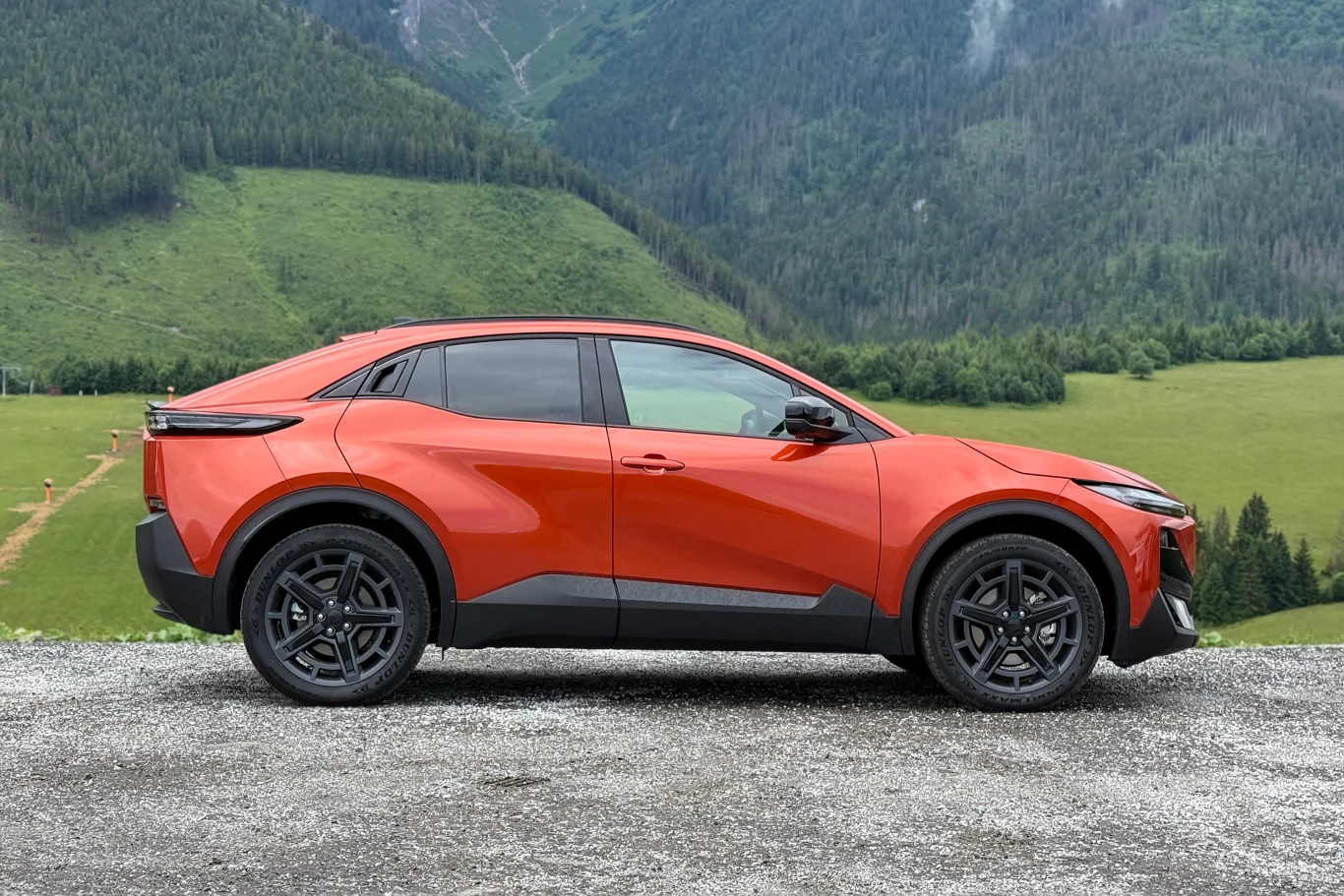 Stylistycznie Subaru Uncharted i Toyota C-HR+ są do siebie bardzo podobne. Czerwony nowoczesny crossover zaparkowany na żwirowej nawierzchni na tle zielonych wzgórz i górzystego krajobrazu.