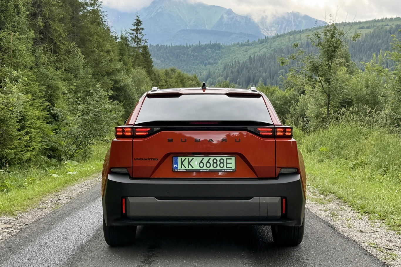 Subaru Uncharted idealnie nadaje się jako kompan do dalszych podróży. Pomarańczowy samochód elektryczny Subaru Solterra stoi na wąskiej asfaltowej drodze otoczonej zielonym lasem, w tle widoczne górskie szczyty i zachmurzone niebo.
