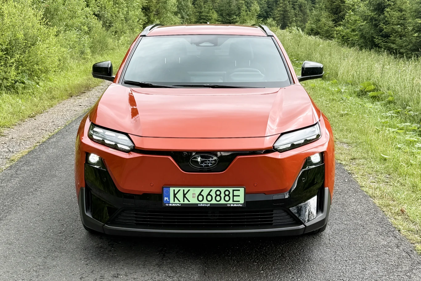Subaru Uncharted Platinum AWD wyposażono w akumulator o pojemności 77 kWh oraz dwa silniki elektryczne. Pomarańczowy samochód elektryczny Subaru prezentuje się na wąskiej, asfaltowej drodze otoczonej zielenią, z widocznym nowoczesnym przodem i zieloną tablicą rejestracyjną.