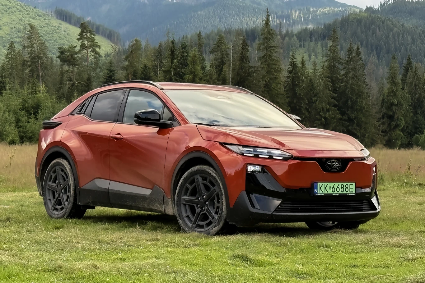Zarówno lekki, jak i cięższy teren, nie stanowi dla Subaru Uncharted najmniejszego wyzwania. Czerwony elektryczny SUV o nowoczesnej sylwetce stoi na trawie na tle gór i lasu.