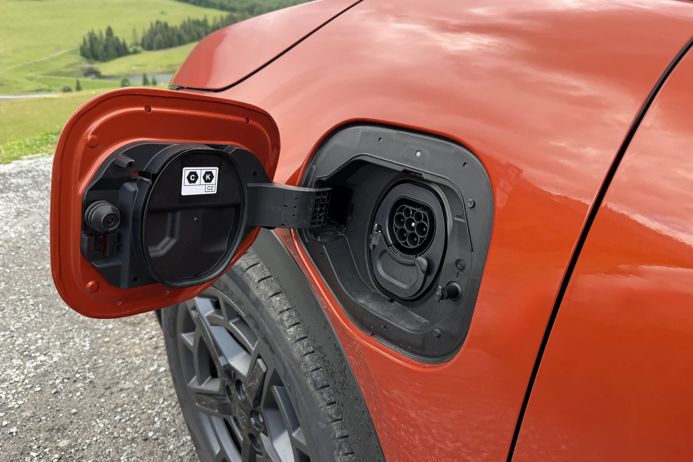 Subaru Uncharted oferuje ładowanie prądem stałym DC z mocą do 150 kW oraz pokładową ładowarkę AC o mocy 22 kW. otwarty wlew ładowania samochodu elektrycznego z widocznym gniazdem podłączania kabla na tle pomarańczowej karoserii i szarego asfaltu