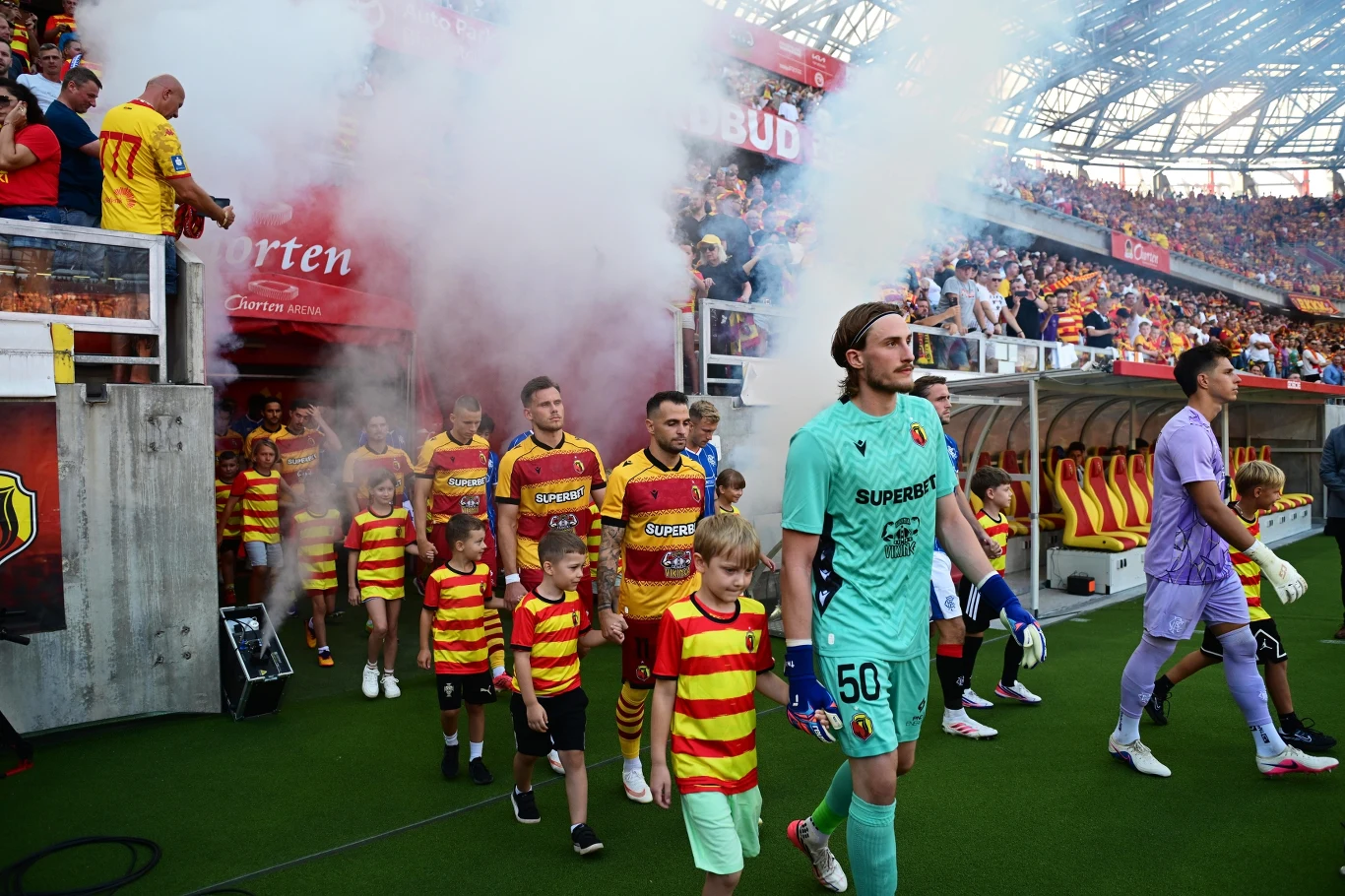 Piłkarze w żółto-czerwonych strojach wychodzą na murawę stadionu w towarzystwie dzieci, z tunelu unosi się biały dym, a na trybunach widać kibiców w klubowych barwach.