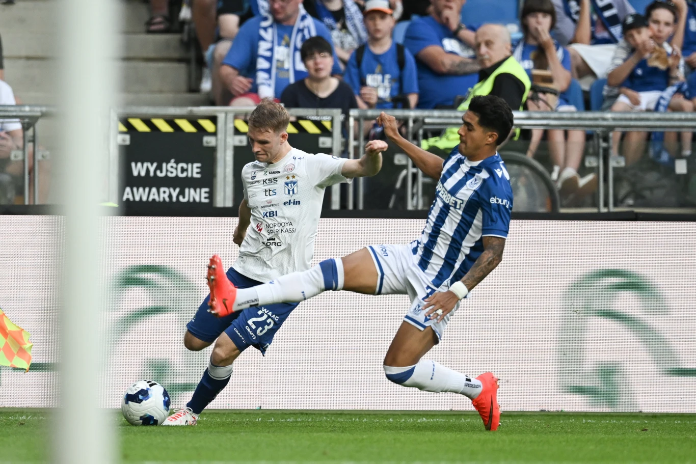 Dwóch piłkarzy walczy o piłkę tuż przy linii bocznej boiska, jeden z nich wślizgiem próbuje odebrać futbolówkę przeciwnikowi; na trybunach widoczna grupa kibiców w niebiesko-białych barwach.