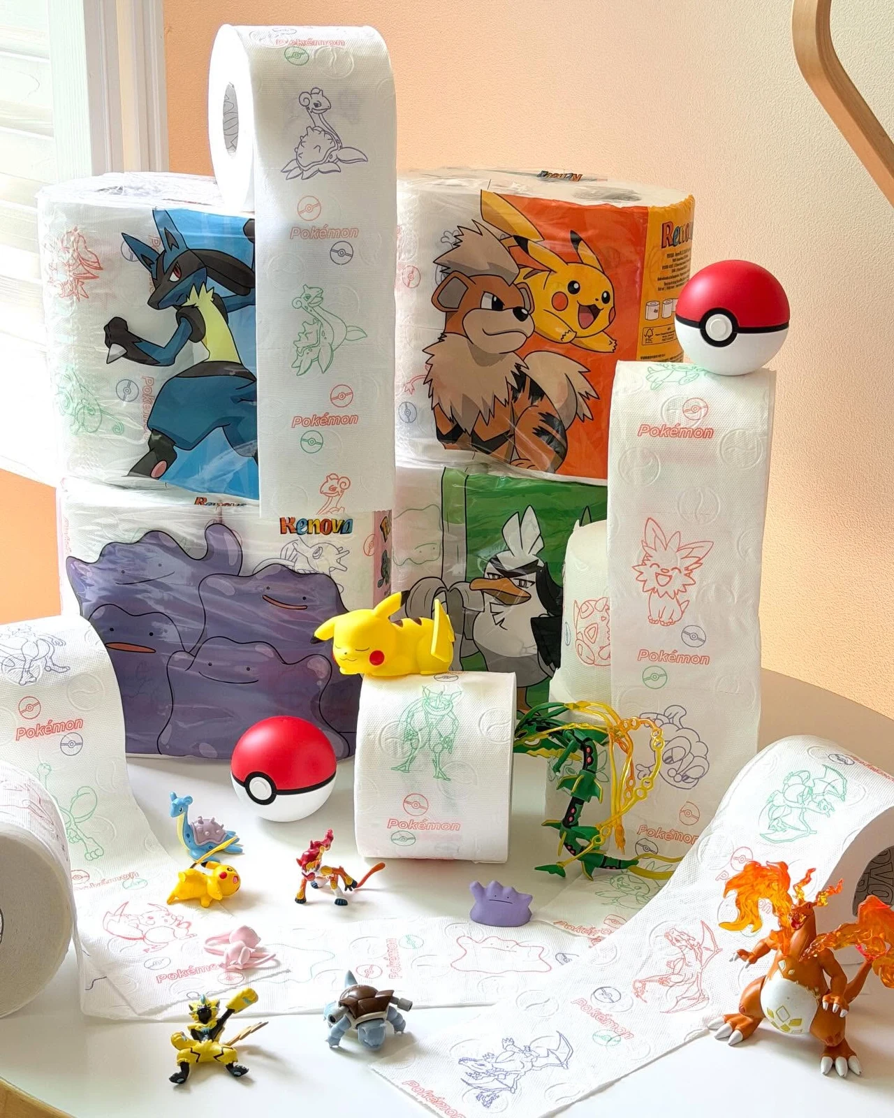 Kolorowe opakowania papieru toaletowego z motywami Pokémon, rozwinięte rolki z nadrukami postaci takich jak Pikachu czy Eevee, plastikowe figurki Pokémonów oraz pokeballe rozłożone wokół produktów.