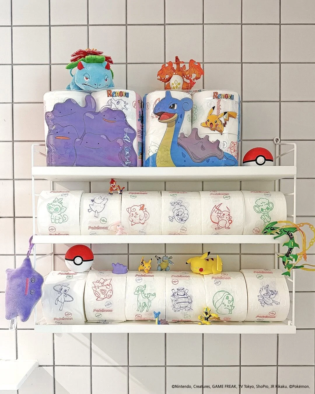 Kolorowe rolki papieru toaletowego z motywami postaci Pokémon, ustawione na białym, metalowym regale, z maskotkami i dekoracjami nawiązującymi do tej popularnej serii gier i animacji.